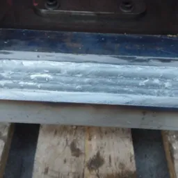 Szczegółowe ujęcie spawu na metalowej konstrukcji, widoczne liczne ściegi spawalnicze nałożone warstwami, spoczywające na drewnianej powierzchni.