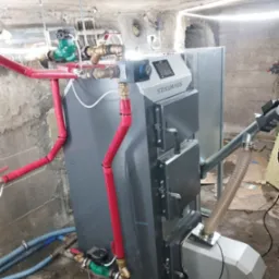 Szary piec CO marki Stalmark z podajnikiem, podłączony do instalacji z rurami w czerwonej izolacji, w piwnicy z surowym betonowym sufitem.