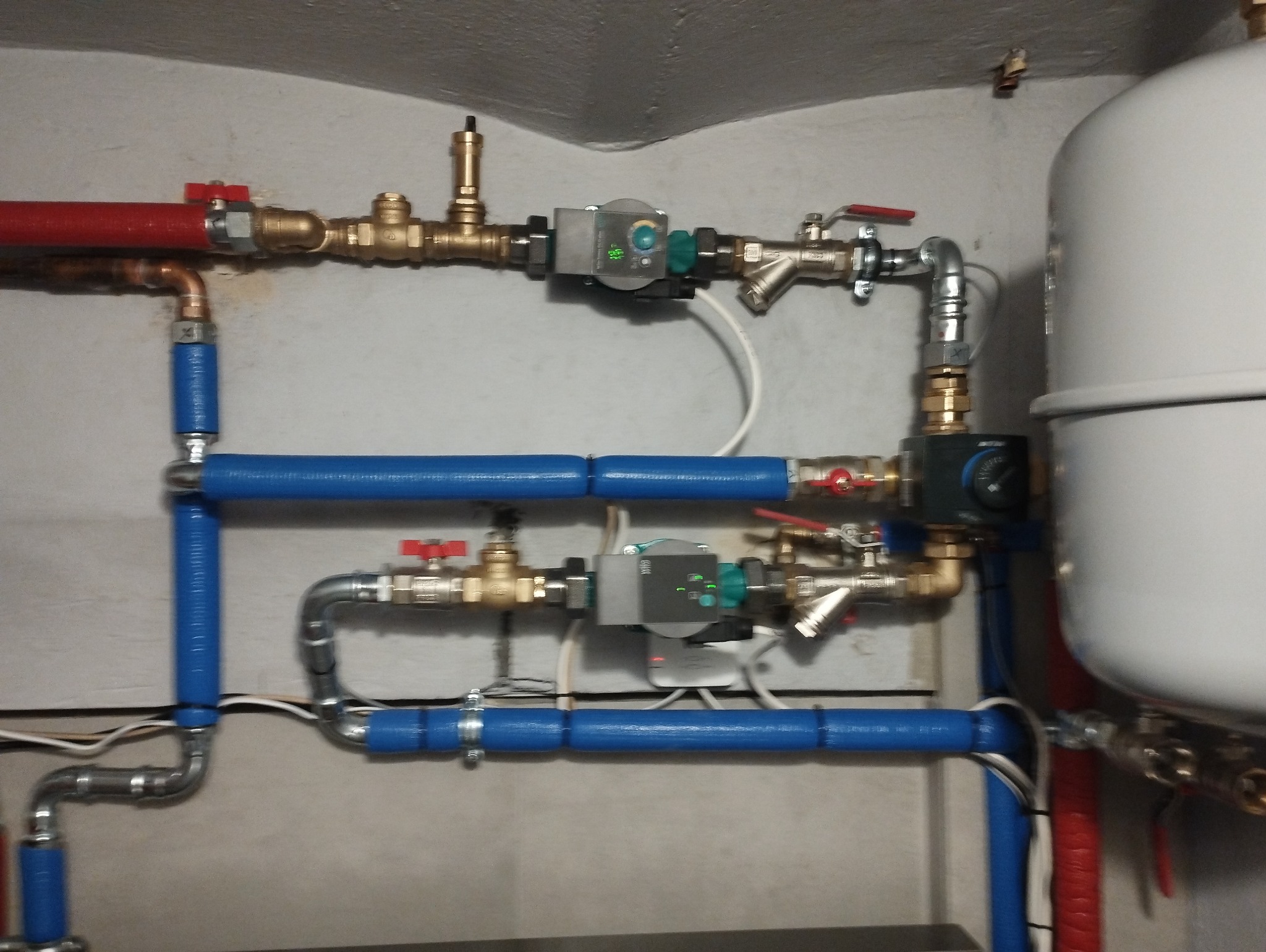 Rozbudowana instalacja hydrauliczna i grzewcza z licznymi rurami w kolorach niebieskim, czerwonym i miedzianym, zaworami, pompami obiegowymi i zbiornikiem wyrównawczym, zamontowana na szarej ścianie.