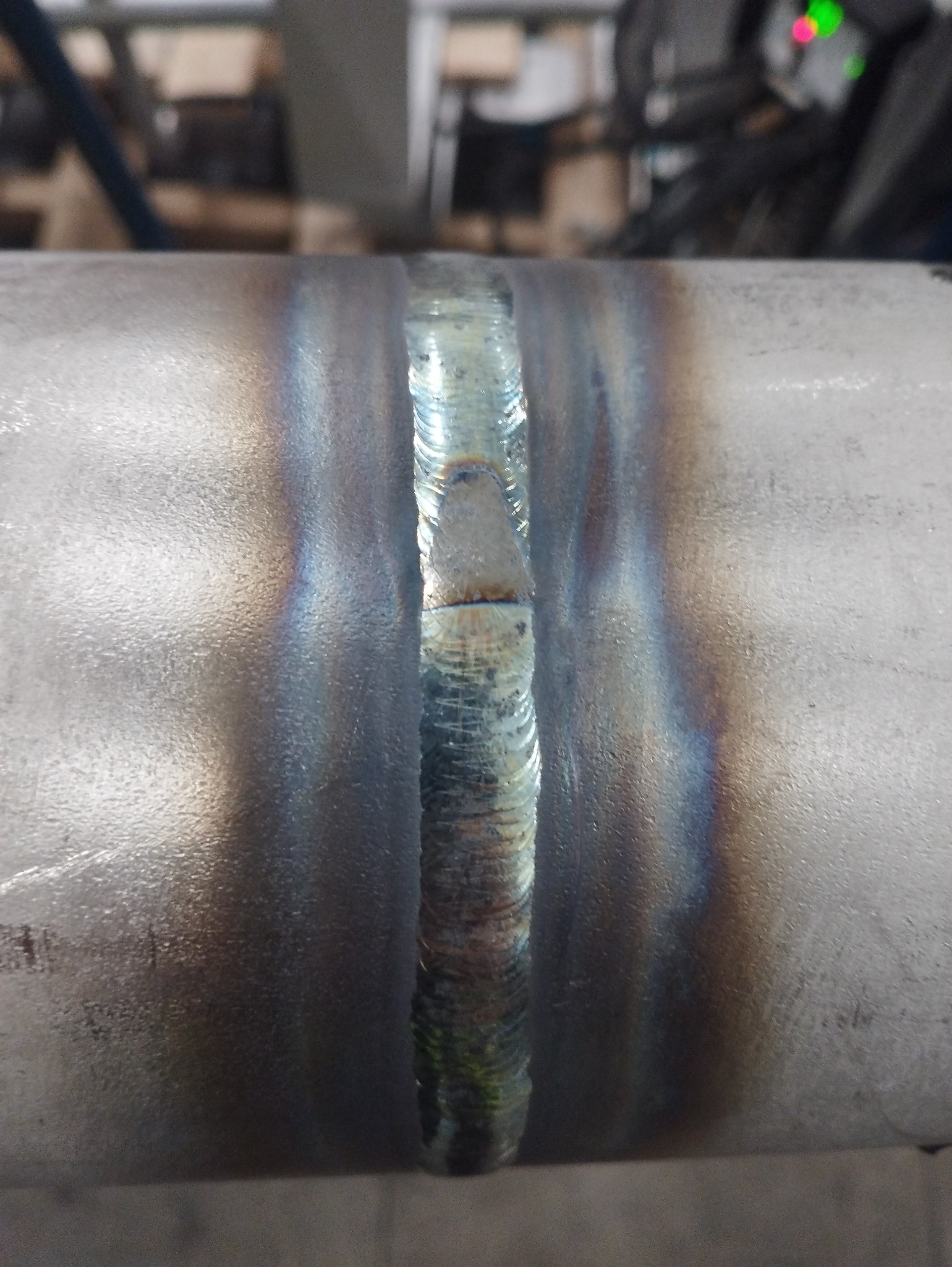 Szlifowany spaw na rurze stalowej, widoczne ślady obróbki i przebarwienia termiczne, w tle zamazane elementy wyposażenia warsztatu.
