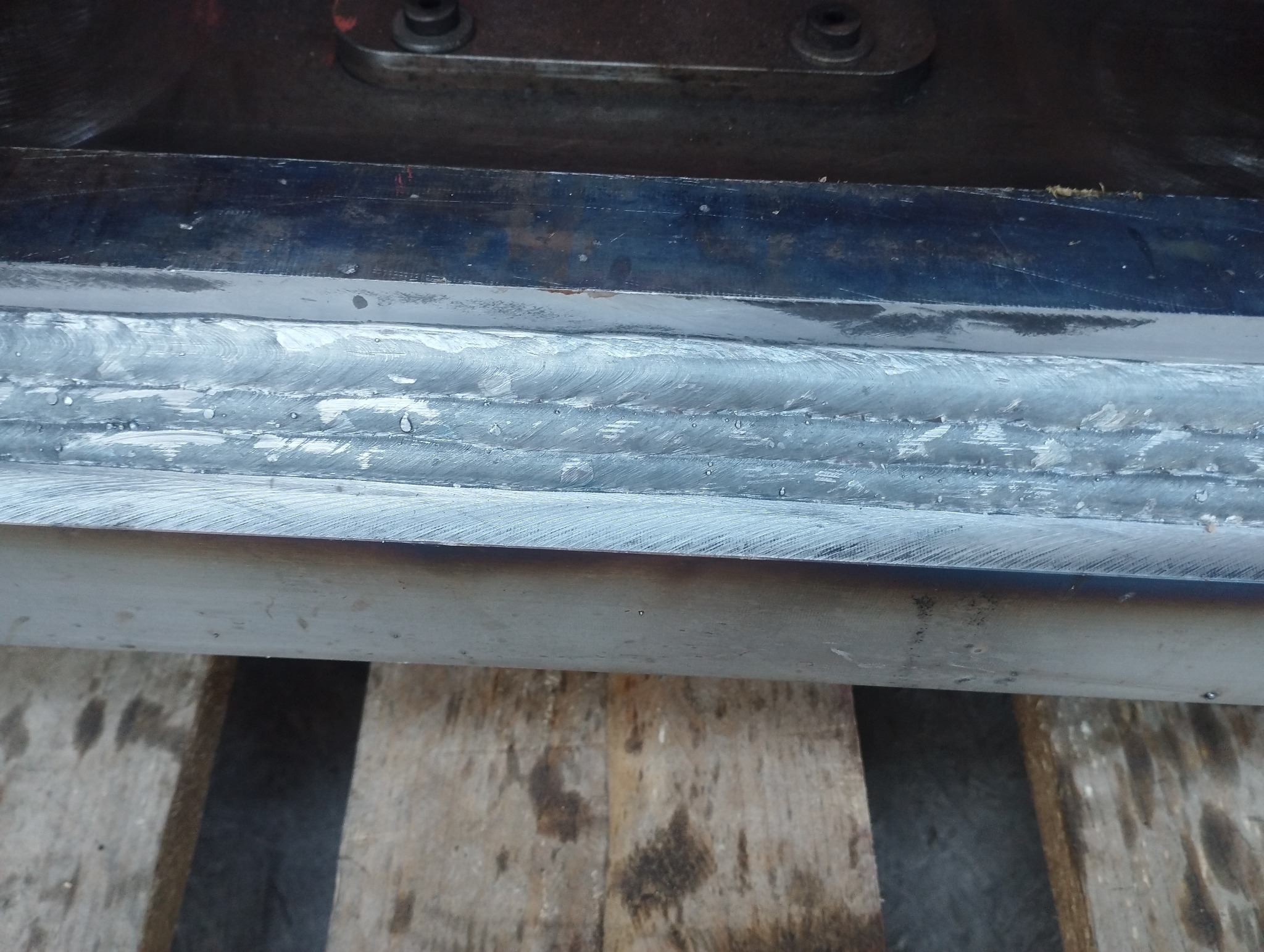 Szczegółowe ujęcie spawu na metalowej konstrukcji, widoczne liczne ściegi spawalnicze nałożone warstwami, spoczywające na drewnianej powierzchni.