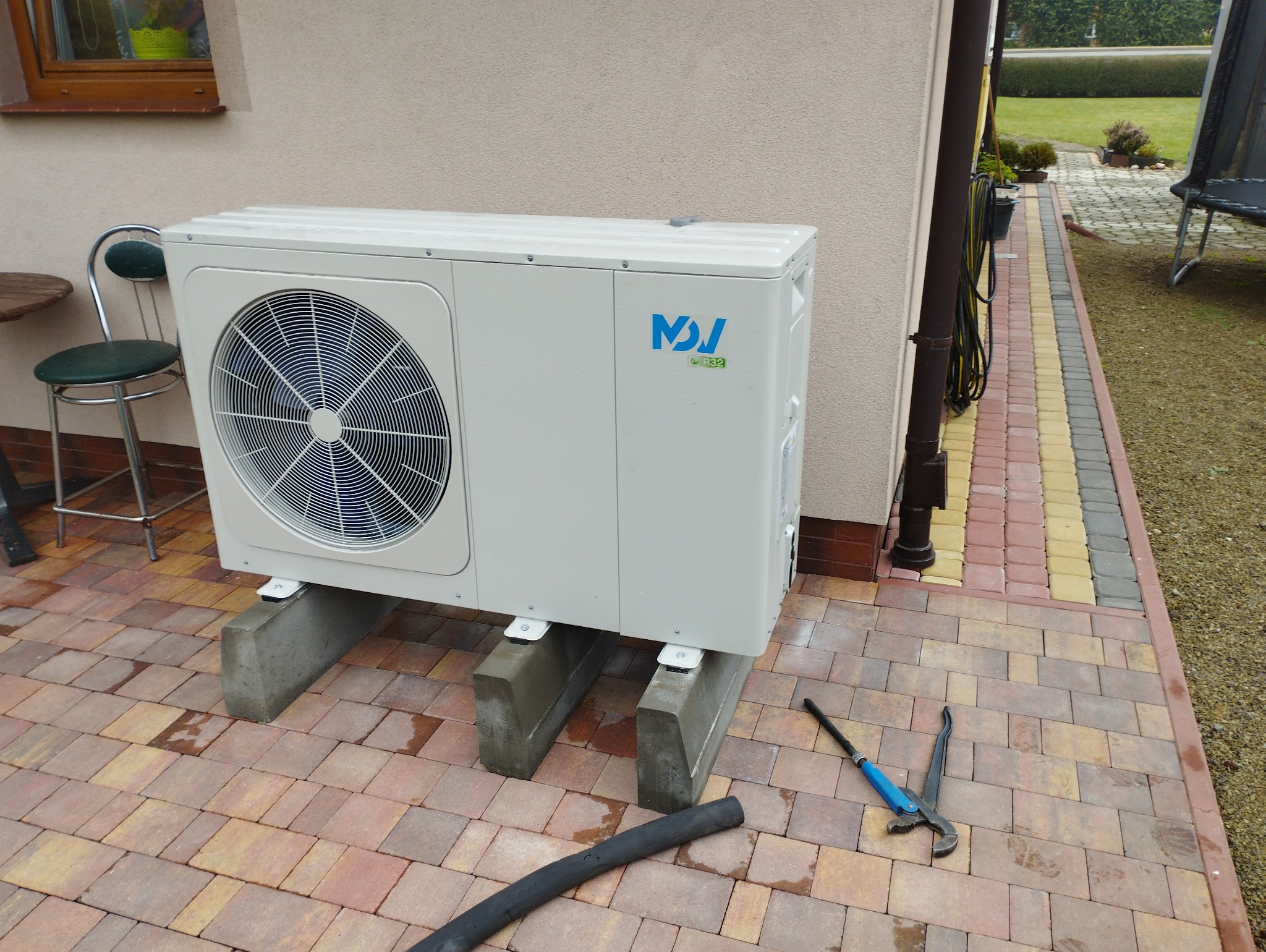 Biała jednostka zewnętrzna klimatyzacji na betonowych wspornikach, z widocznymi narzędziami hydraulicznymi na kostce brukowej.