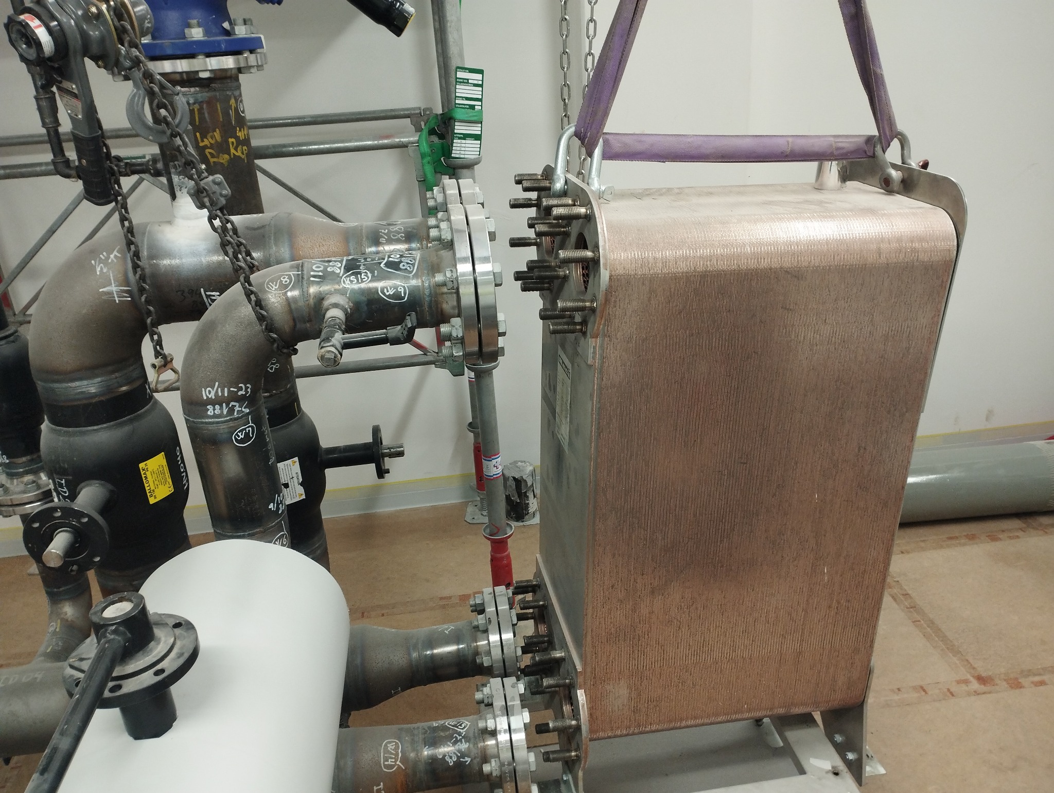 Wymiennik ciepła płytowy, podwieszony na pasach, w trakcie montażu do instalacji rurociągów stalowych z licznymi złączami kołnierzowymi i oznaczeniami spawalniczymi.