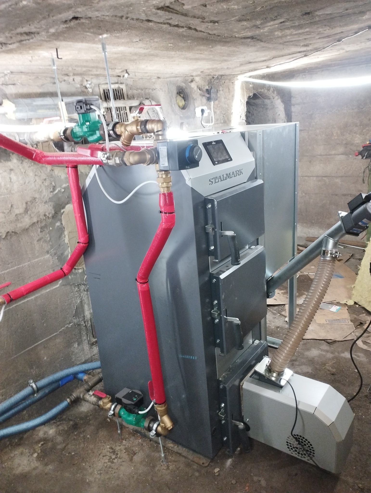 Szary piec CO marki Stalmark z podajnikiem, podłączony do instalacji z rurami w czerwonej izolacji, w piwnicy z surowym betonowym sufitem.