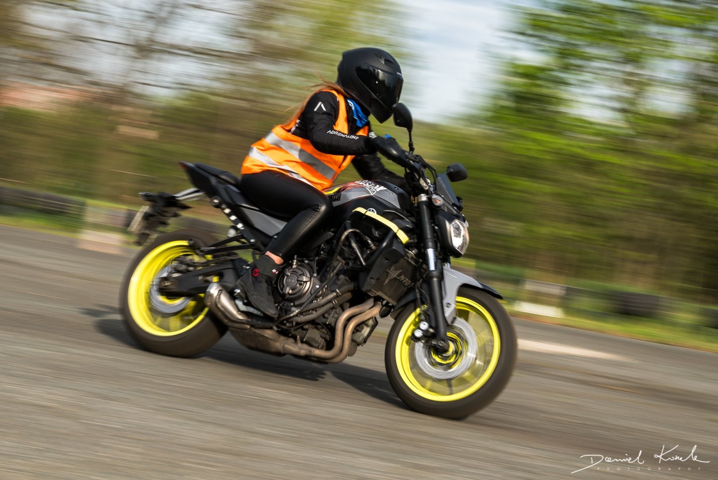 Kobieta w kasku i kamizelce odblaskowej na motocyklu Yamaha MT-07 z żółtymi felgami, dynamiczne ujęcie w ruchu na torze.