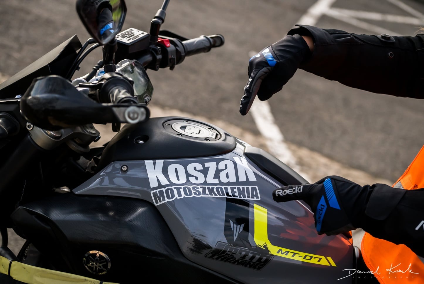 Zbliżenie na motocykl MT-07 z naklejkami 'Koszak Motoszkolenia', instruktor w rękawicach wskazuje na zbiornik paliwa.