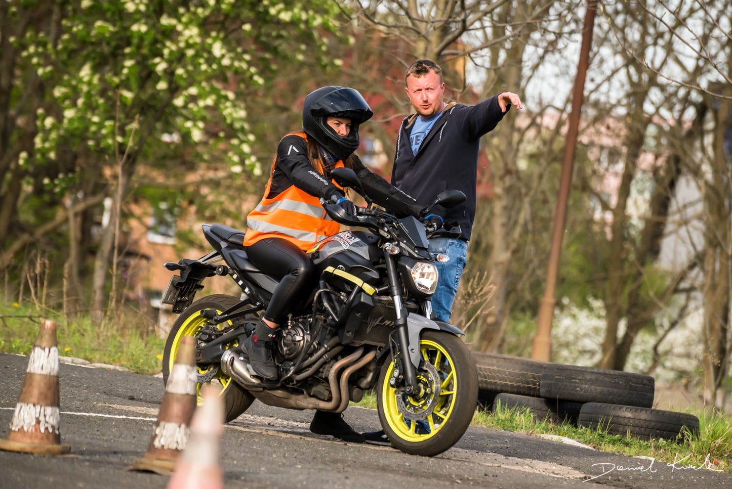 Kobieta w czarnym kasku i pomarańczowej kamizelce na motocyklu Yamaha MT-07 z żółtymi felgami, instruktor wskazuje kierunek jazdy na placu manewrowym z pachołkami i oponami.