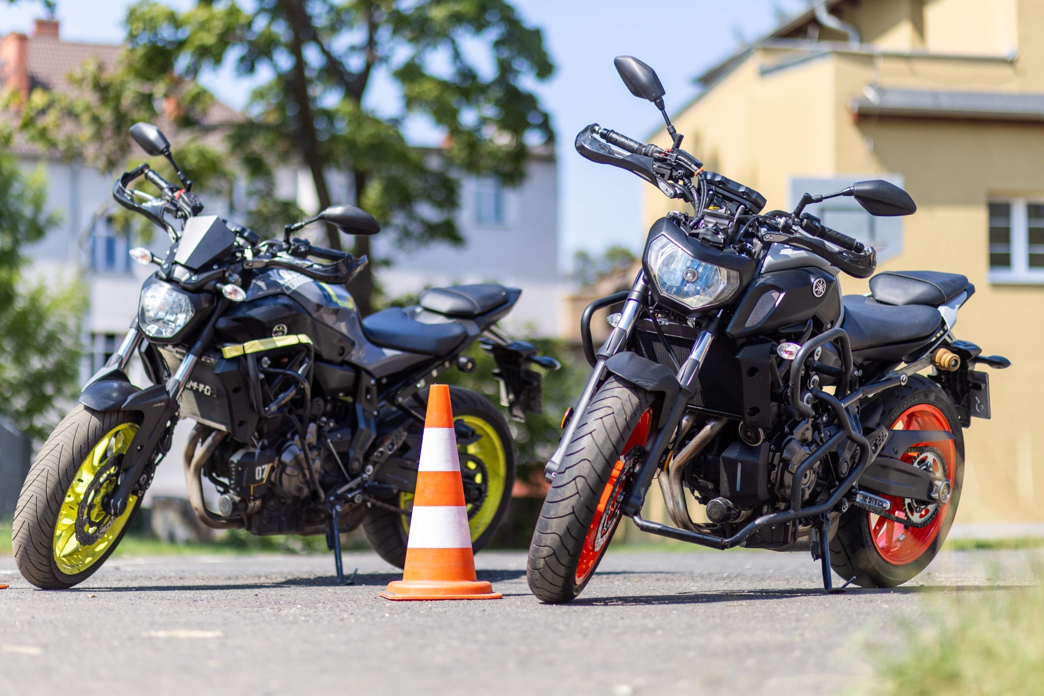 Dwa motocykle Yamaha MT-07, jeden z żółtymi felgami, drugi z pomarańczowymi, stojące na asfalcie obok pomarańczowo-białego pachołka treningowego, w tle budynki.