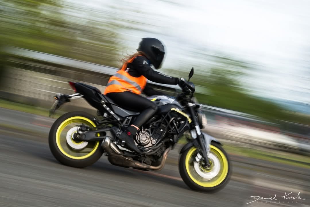 Kobieta w czarnym kasku i pomarańczowej kamizelce odblaskowej na czarno-szarym motocyklu z żółtymi felgami w dynamicznym ujęciu z efektem rozmycia ruchu.