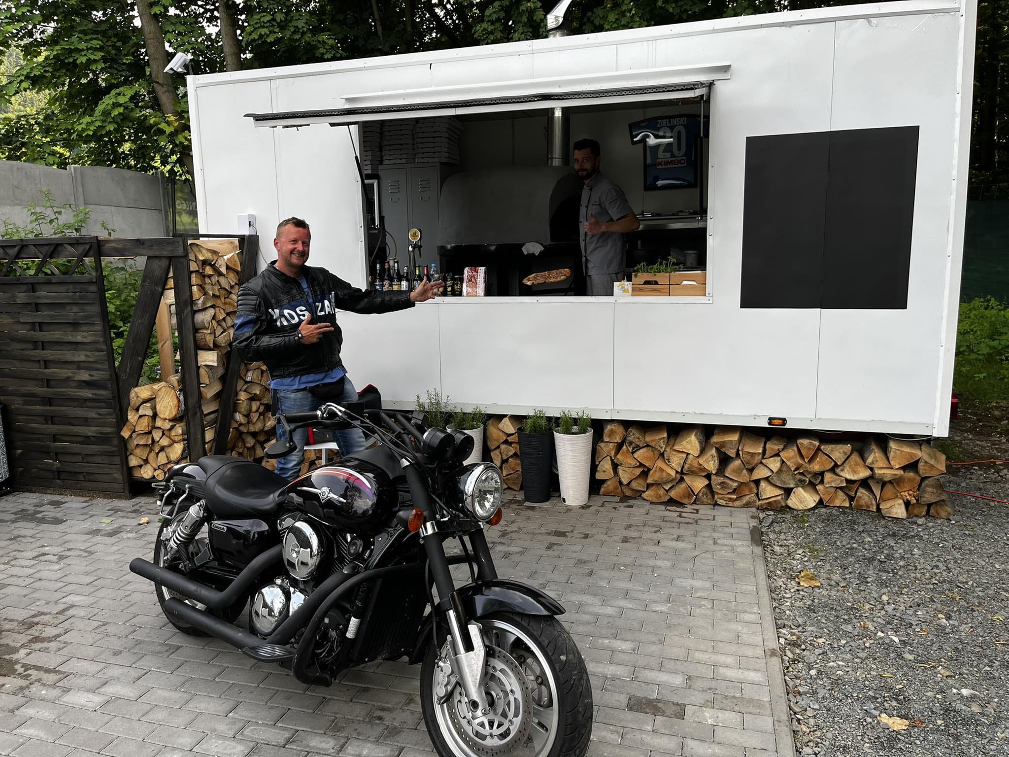 Mobilna pizzeria z piecem opalanym drewnem, motocykl na pierwszym planie, mężczyzna w skórzanej kurtce gestykuluje, w tle kucharz przy piecu, na ścianie koszulka piłkarska z nazwiskiem Zieliński.