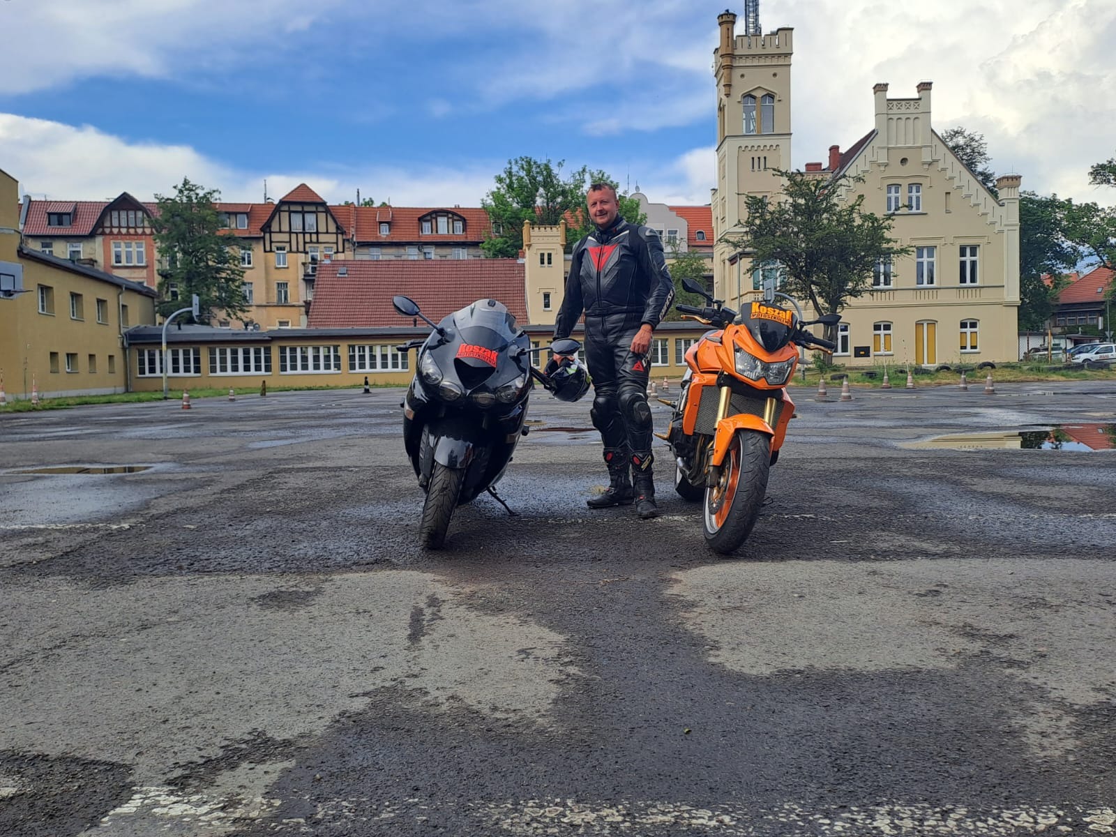 Instruktor w skórzanym kombinezonie stoi między czarnym i pomarańczowym motocyklem na placu manewrowym szkoły jazdy, w tle budynek z wieżą.