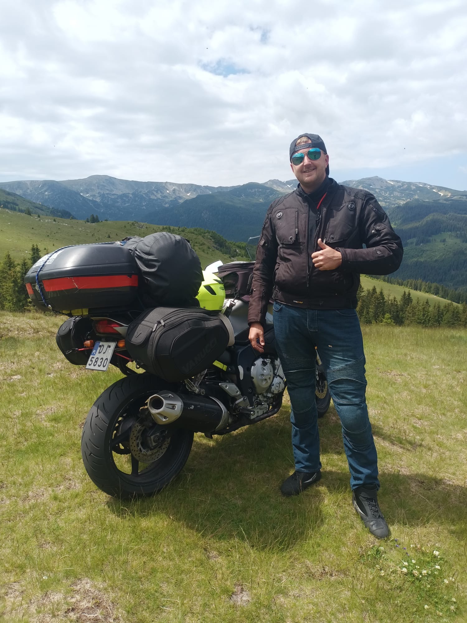 Mężczyzna w kurtce motocyklowej i dżinsach, kciuk w górę, stoi obok motocykla turystycznego z bagażami na tle górskiego krajobrazu.