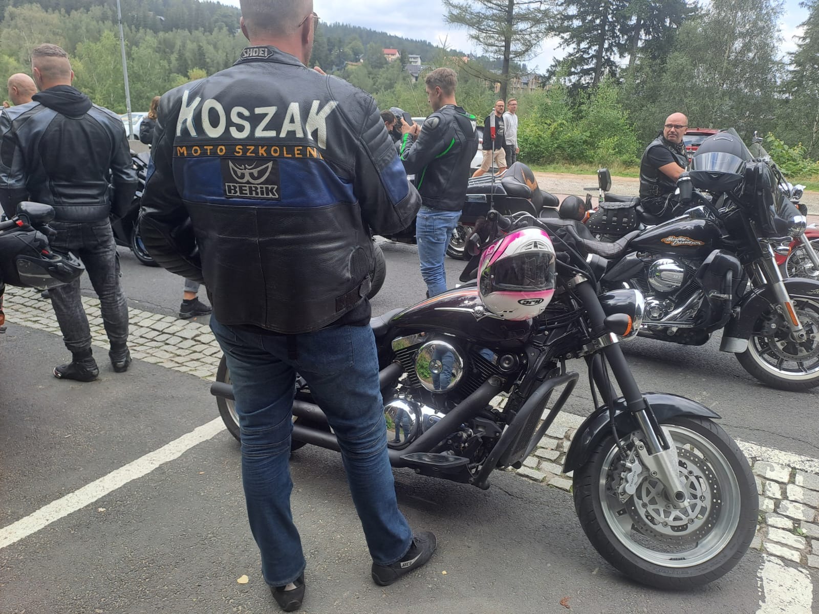 Grupa motocyklistów w skórzanych kurtkach stoi obok zaparkowanych motocykli, na jednym z nich biało-różowy kask.