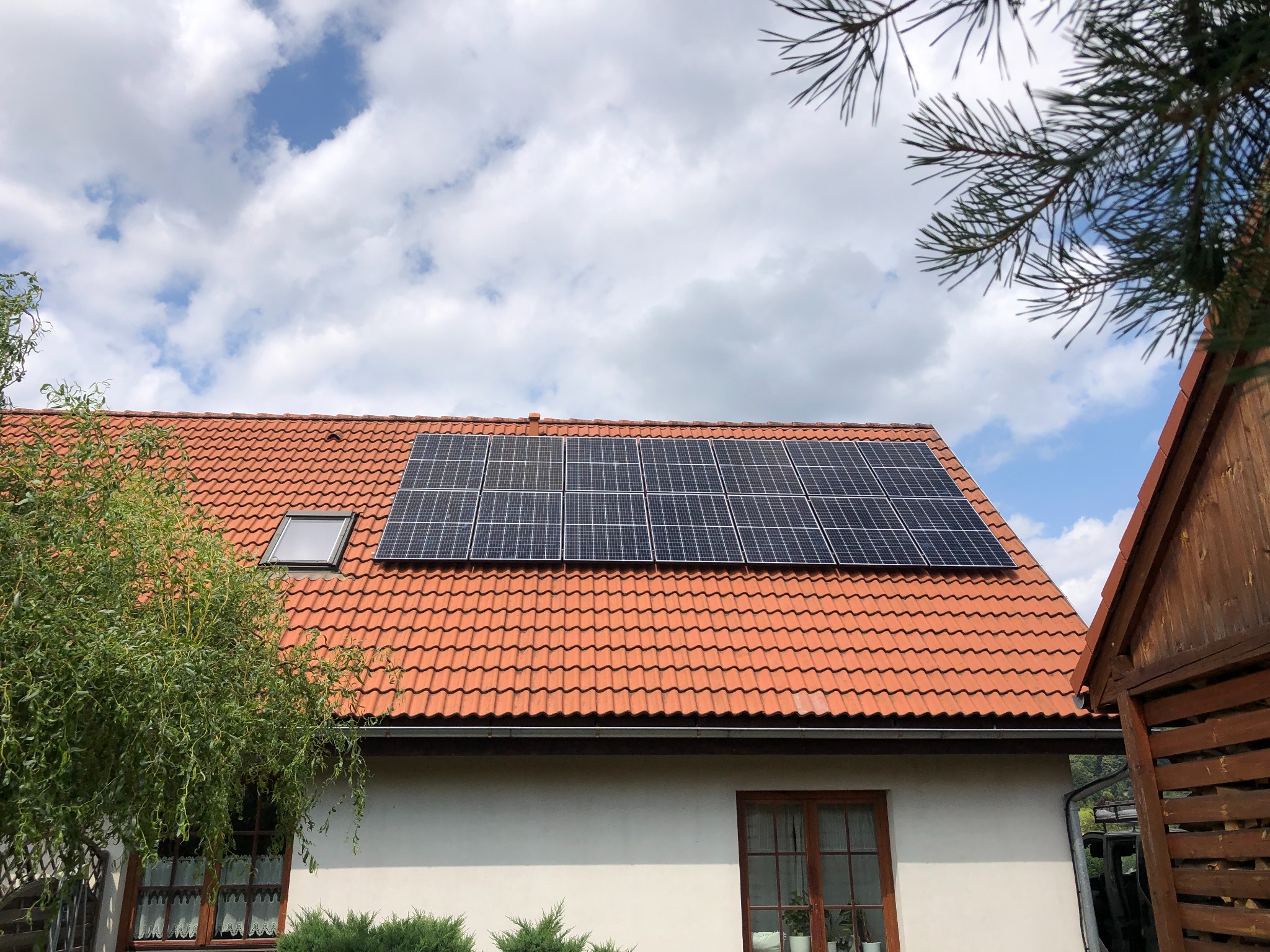Panele fotowoltaiczne na dachu domu z czerwoną dachówką, widok z poziomu ziemi, z elementami zieleni w kadrze.