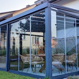 E-TECH DANIEL KOTLIŃSKI - Zadaszony taras z metalową konstrukcją i szklanymi ścianami przesuwnymi. Wewnątrz drewniany stół i wiklinowe krzesła. Widok na ogród i budynki w tle.