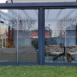 E-TECH DANIEL KOTLIŃSKI - Nowoczesna, szklana zabudowa tarasu z ciemną ramą, widok na meble ogrodowe i zieleń. Przesuwne drzwi zapewniają dostęp do wnętrza, tworząc komfortową przestrzeń.