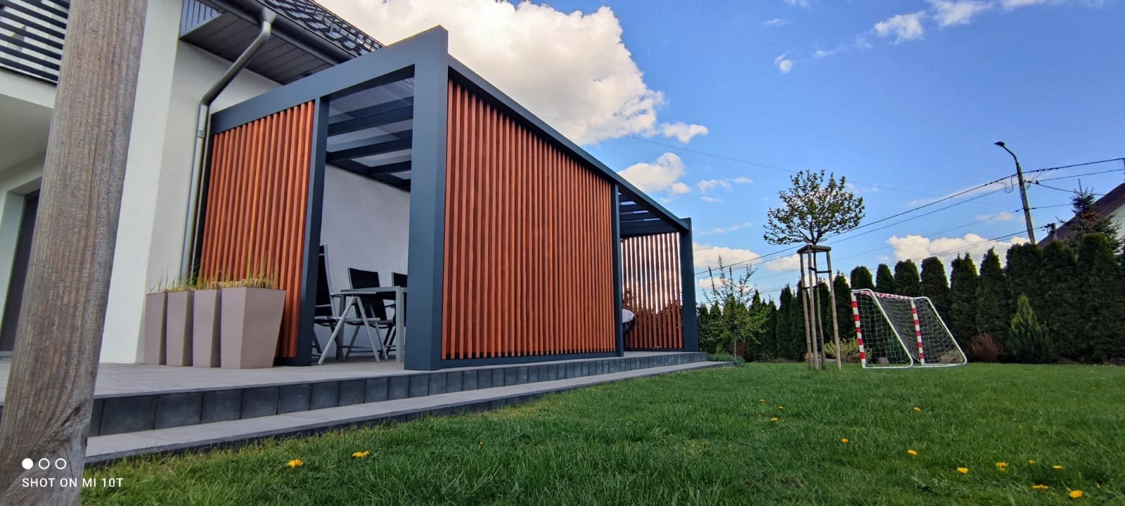 Nowoczesna pergola z drewnianymi panelami i metalową konstrukcją na tle domu i zielonego trawnika. Widoczny stół z krzesłami wewnątrz.