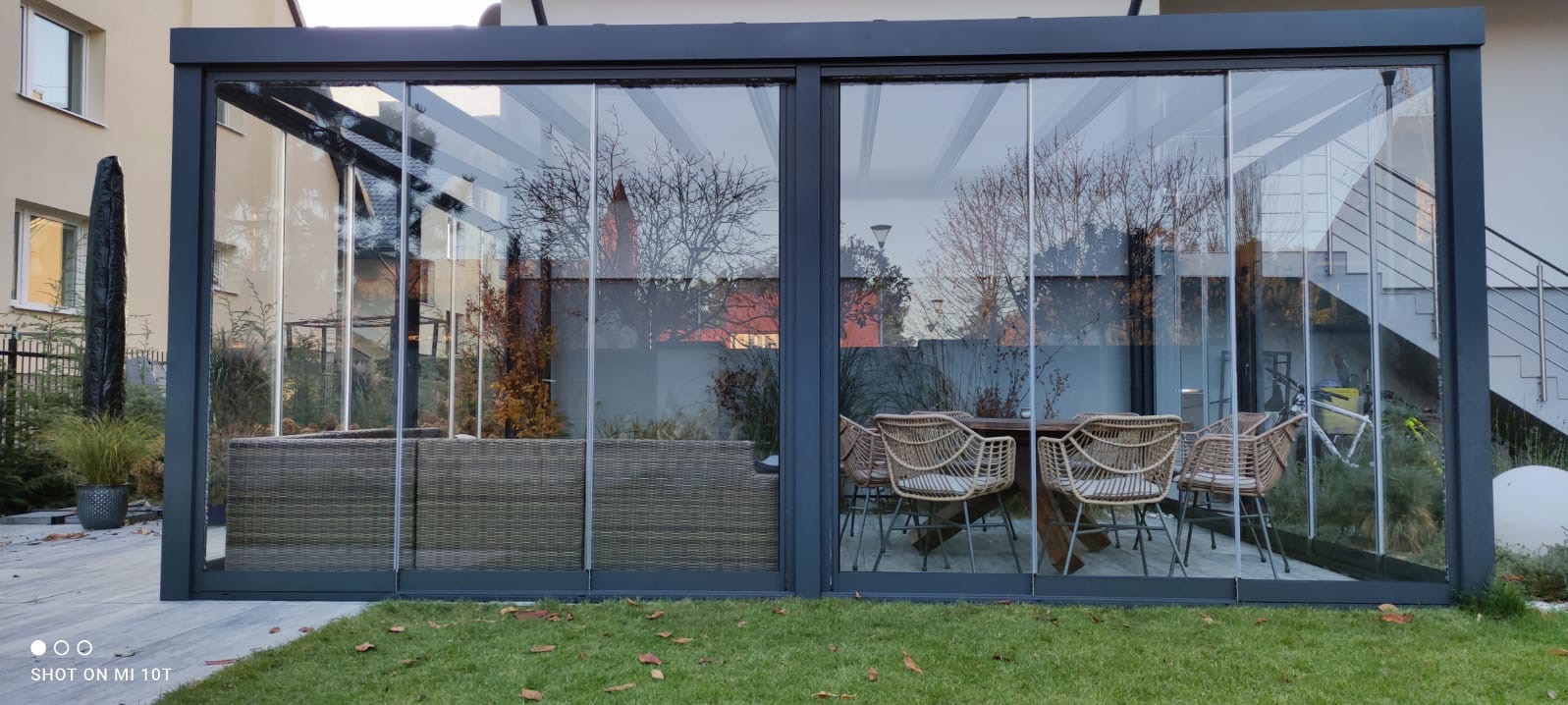 Nowoczesna, szklana zabudowa tarasu z ciemną ramą, widok na meble ogrodowe i zieleń. Przesuwne drzwi zapewniają dostęp do wnętrza, tworząc komfortową przestrzeń.