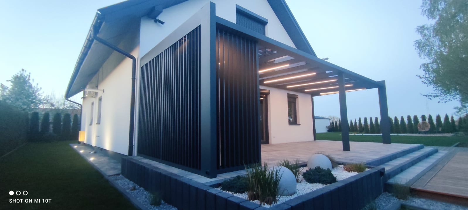 Nowoczesna, antracytowa pergola metalowa z lamelami i oświetleniem LED, przylegająca do jasnego domu z ogrodem. Widok z zewnątrz, wieczorne oświetlenie.