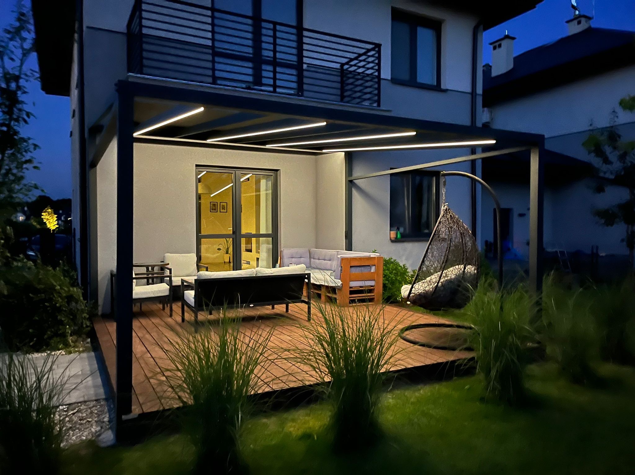 Nowoczesna, oświetlona pergola z drewnianym tarasem i meblami ogrodowymi, tworząca przytulną strefę relaksu przy domu. Widok wieczorny.