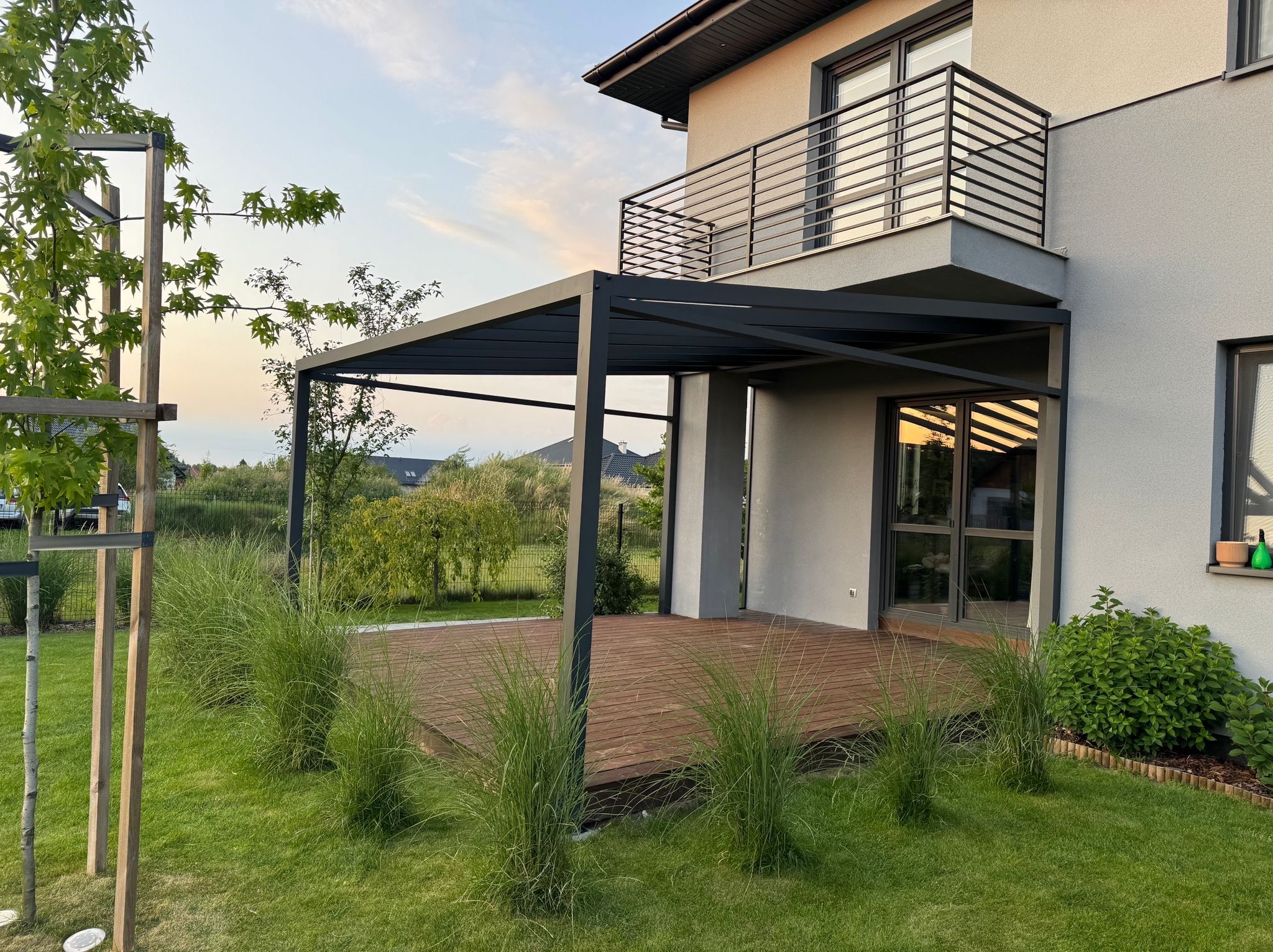Nowoczesna pergola z drewnianym tarasem i ozdobną trawą na tle domu z balkonem. Minimalistyczny design, stonowana kolorystyka, widok na ogród.