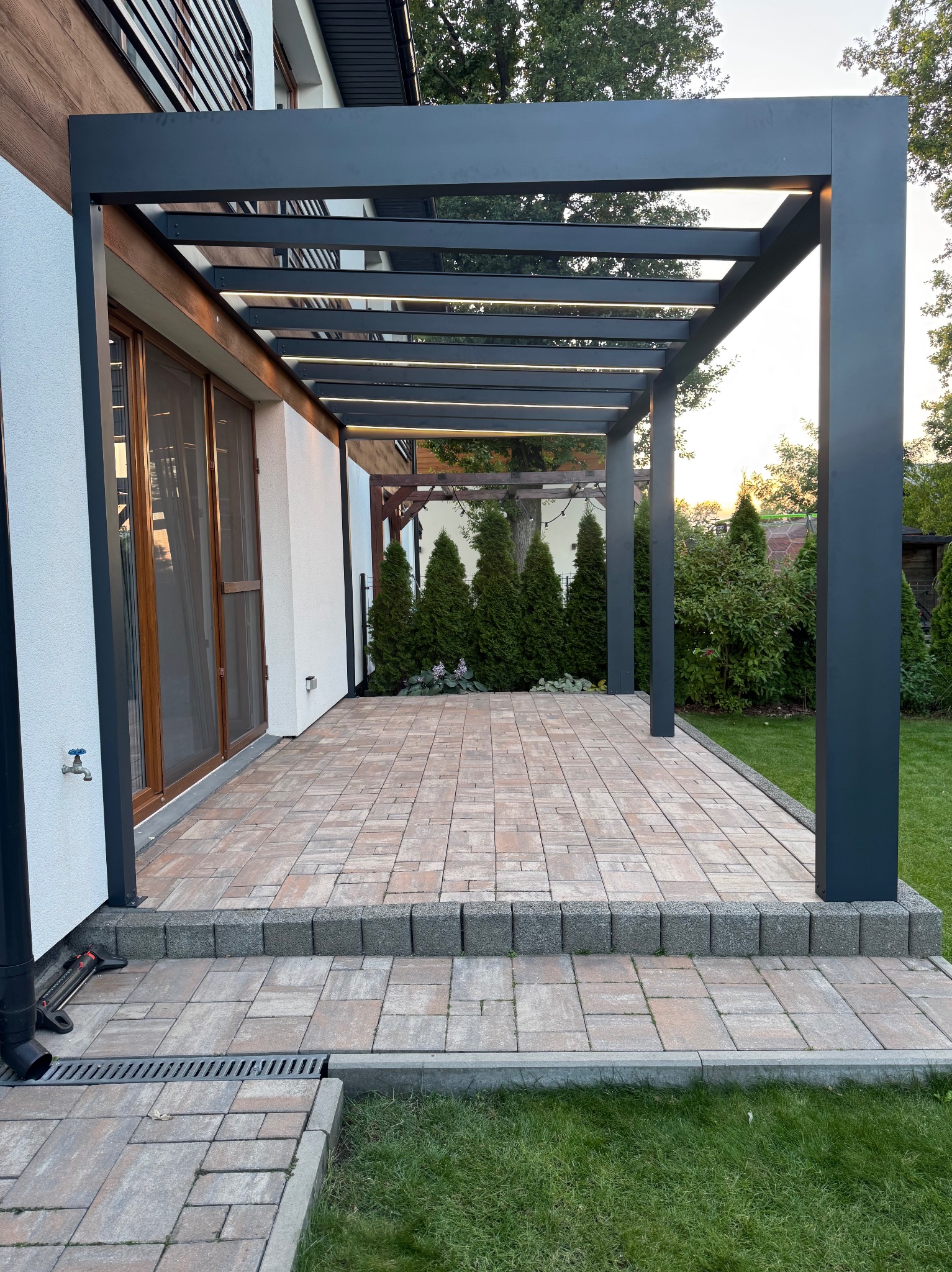 Nowoczesna, grafitowa pergola metalowa z podświetleniem LED, przylegająca do budynku z brukowanym tarasem. Widok na ogród w tle.