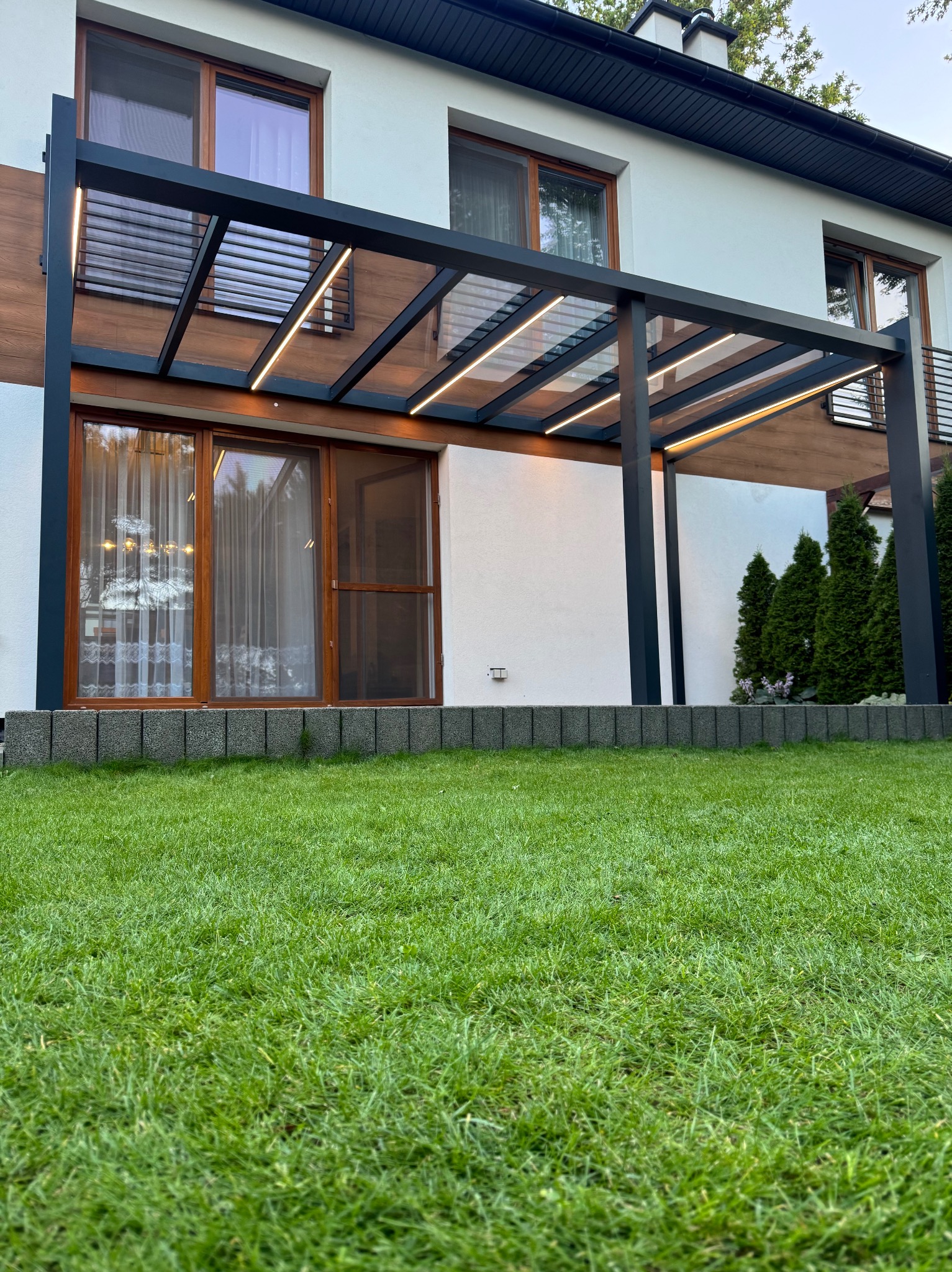 Nowoczesna pergola z oświetleniem LED, przylegająca do budynku z drewnianymi akcentami. Widok z poziomu trawnika, na pierwszym planie soczysta zieleń.