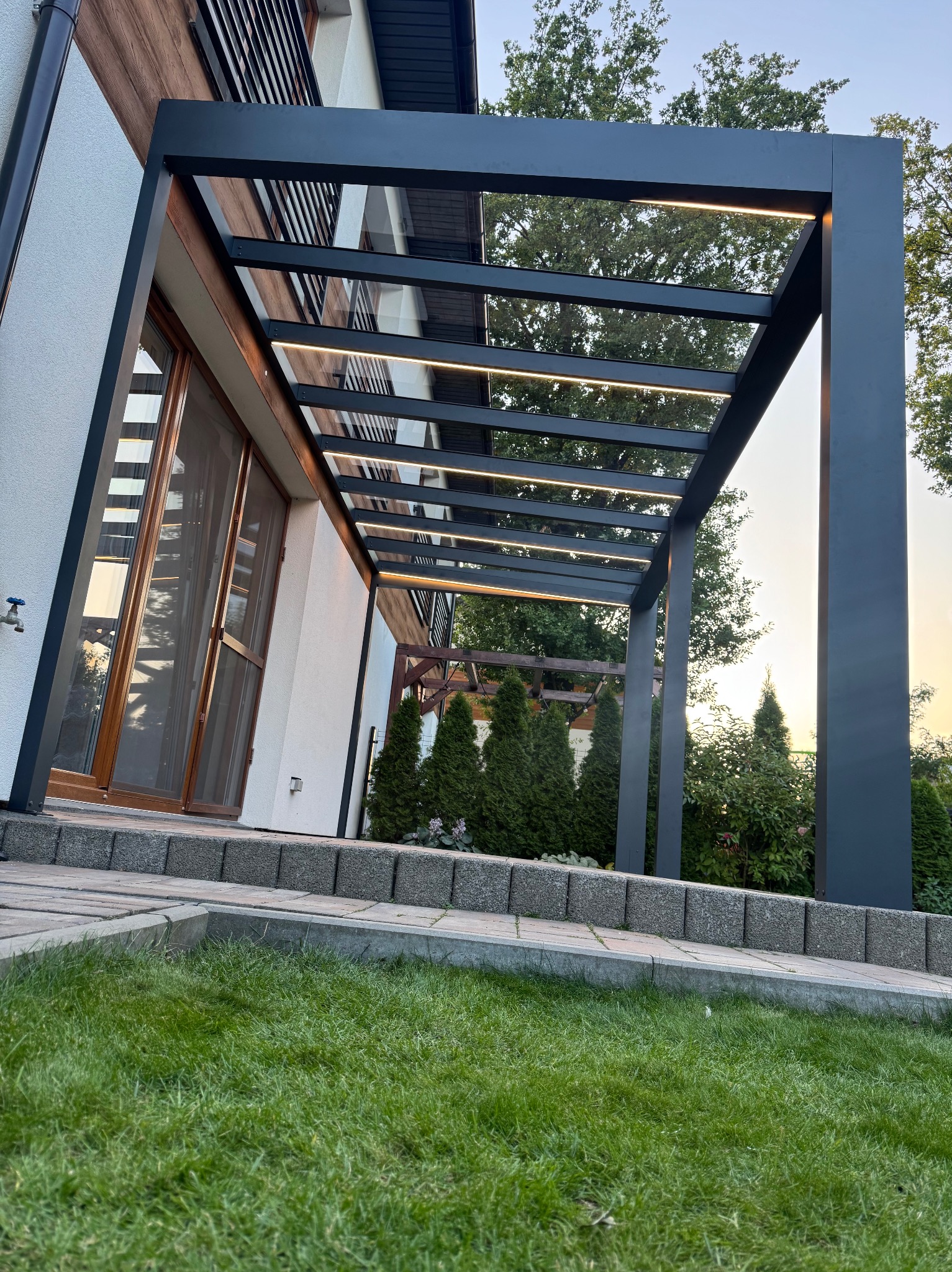 Nowoczesna pergola z oświetleniem LED, przylegająca do domu z drewnianymi elementami, widok z poziomu trawnika. Minimalistyczny design, ciepłe światło.