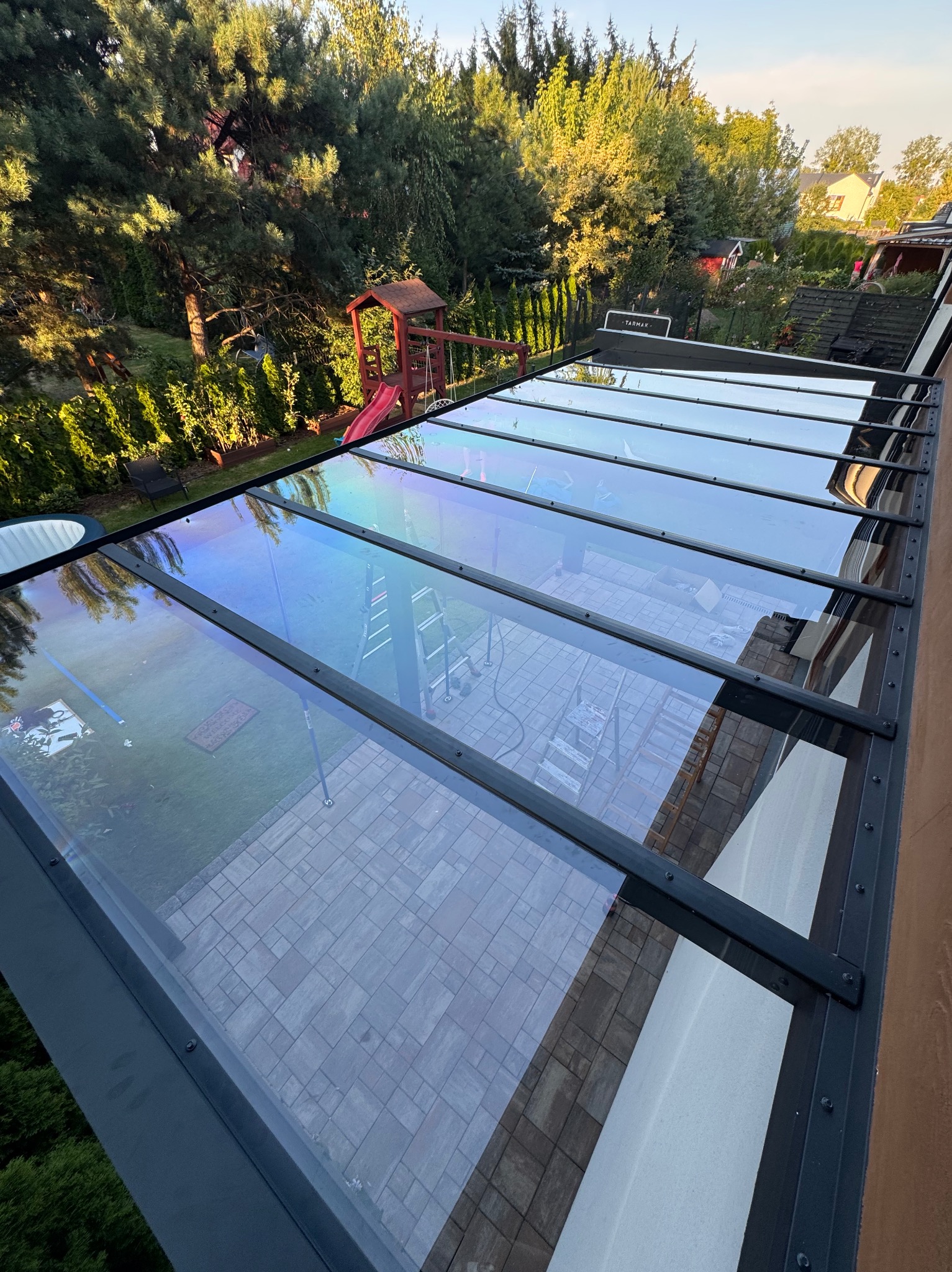 Szklana pergola z czarną, metalową ramą, widok z góry na taras z kostki, zieleń ogrodu w tle. Widoczne odbicie drabiny w szklanej powierzchni.