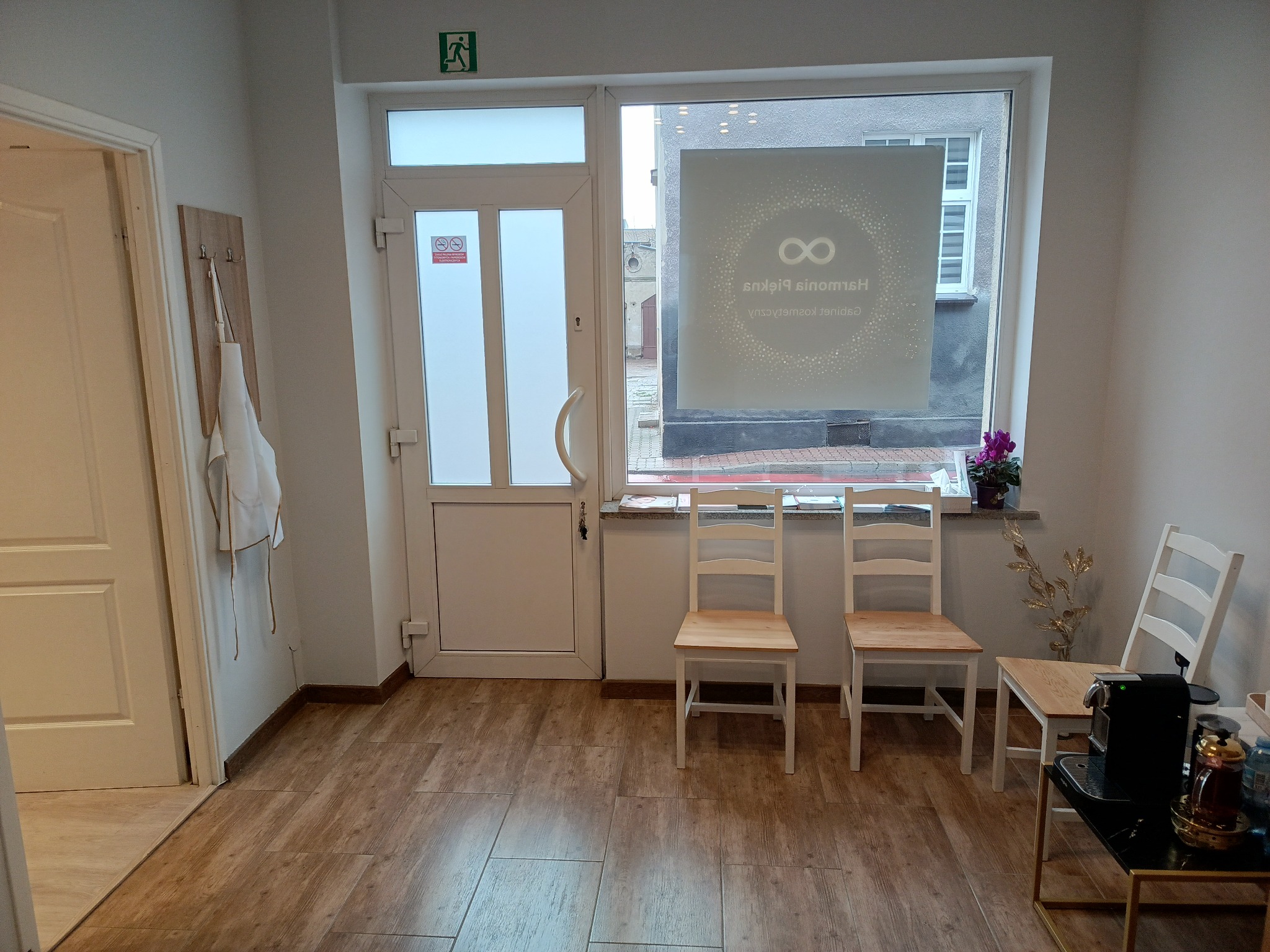 Wnętrze salonu z białymi ścianami, drewnianą podłogą, wejściem z przeszklonymi drzwiami i oknem z naklejonym logo, trzema krzesłami i wieszakiem z fartuchem.