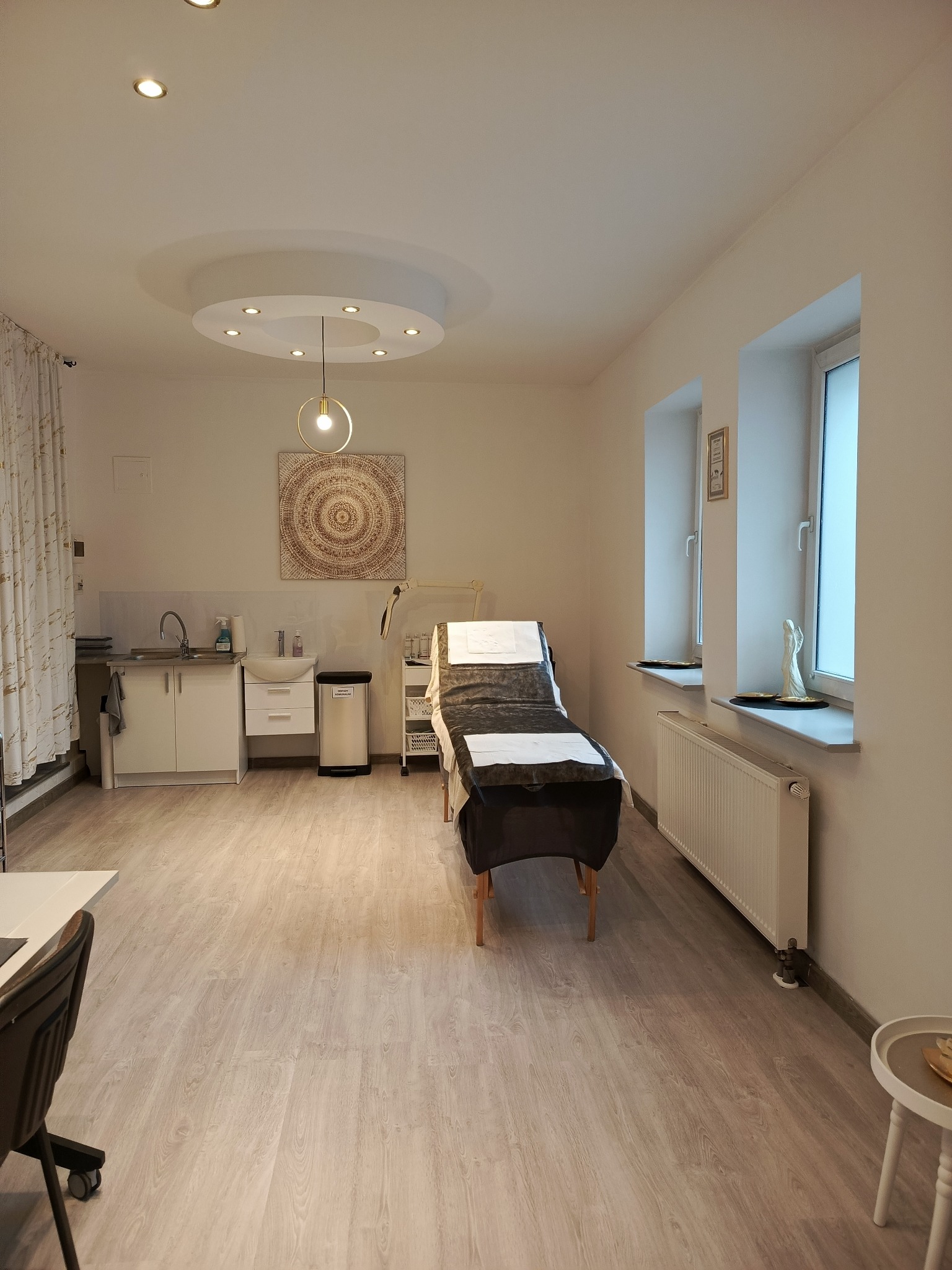 Jasne wnętrze gabinetu kosmetycznego z łóżkiem zabiegowym, umywalką, dekoracyjnym obrazem na ścianie i dwoma oknami z figurkami aniołów na parapetach.