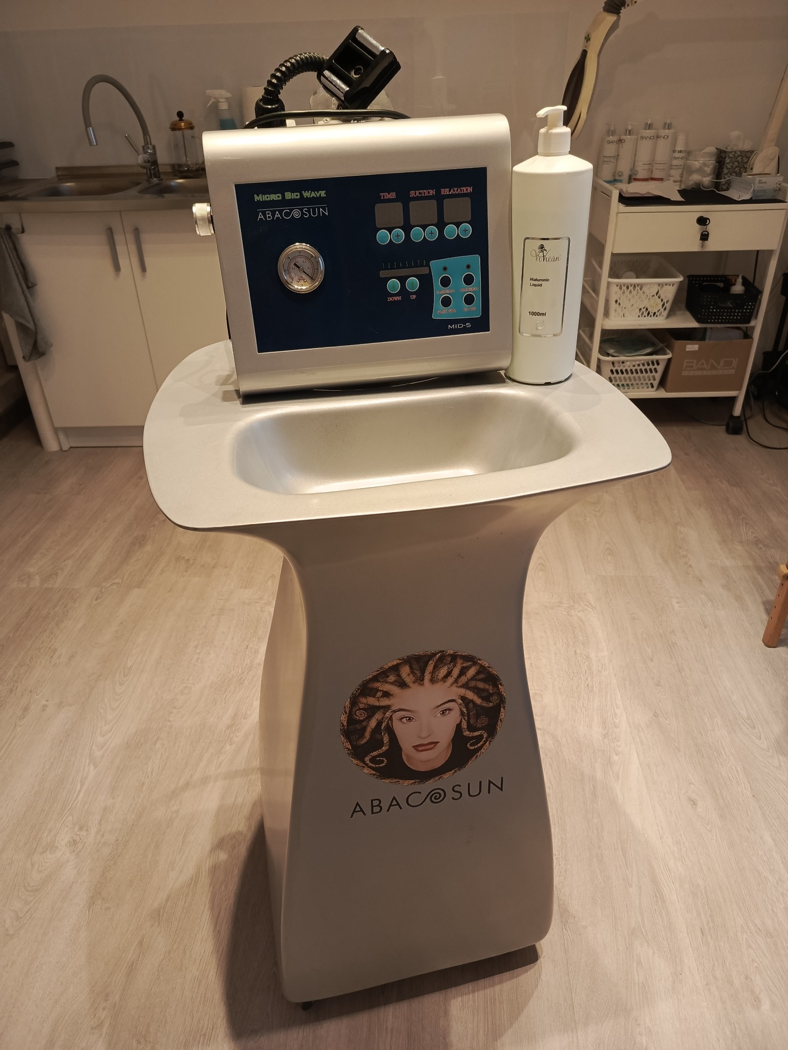 Urządzenie kosmetyczne Micro Bio Wave Abacosun z panelem sterowania, manometrem i butelką płynu Vihean Hyaluronic Liquid 1000ml, stojące na postumencie z wizerunkiem kobiety z dredami, w tle...