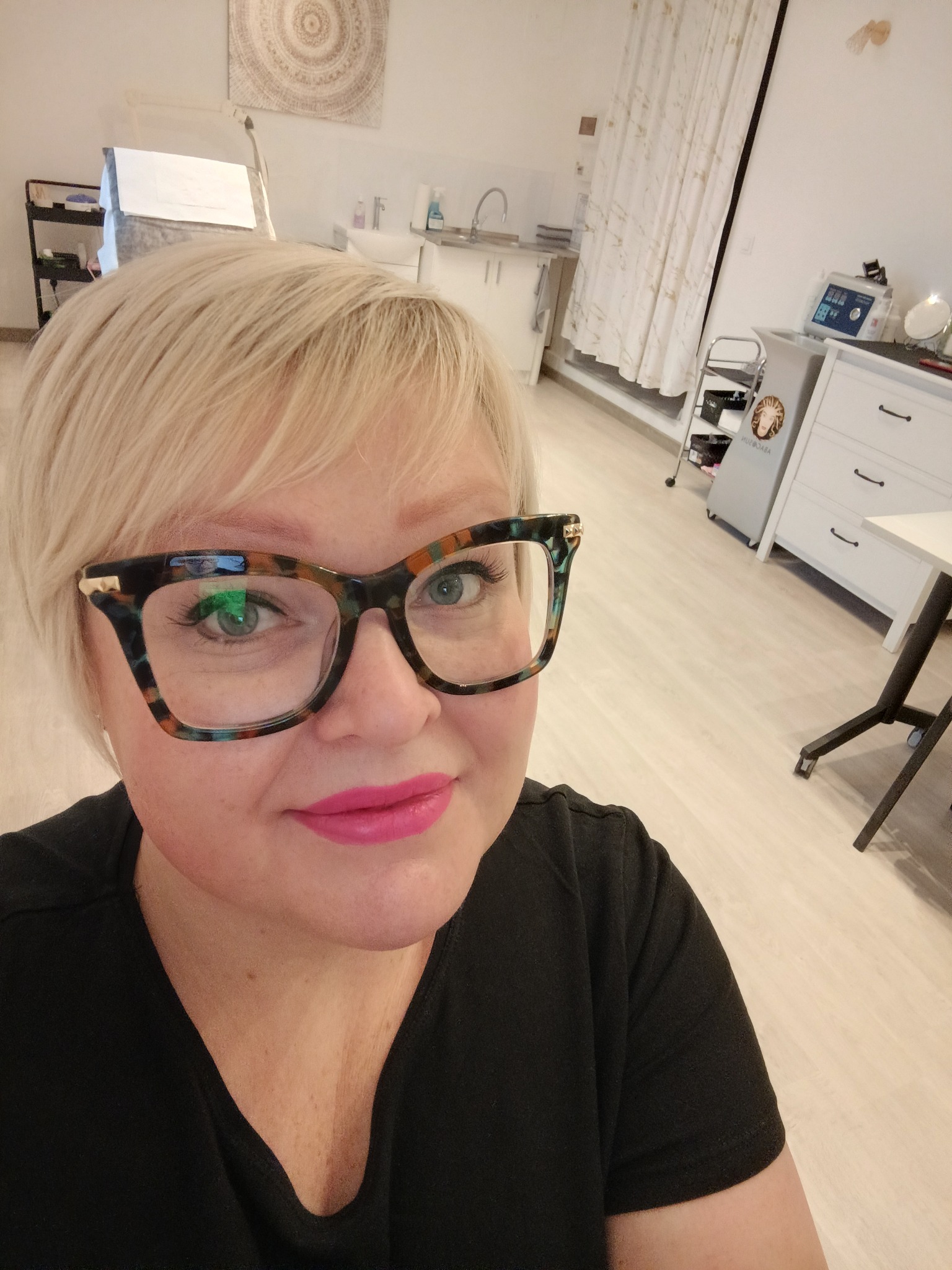 Portret kobiety w okularach z grubymi oprawkami, z mocnym różowym kolorem ust, w tle widoczne wyposażenie gabinetu kosmetycznego.