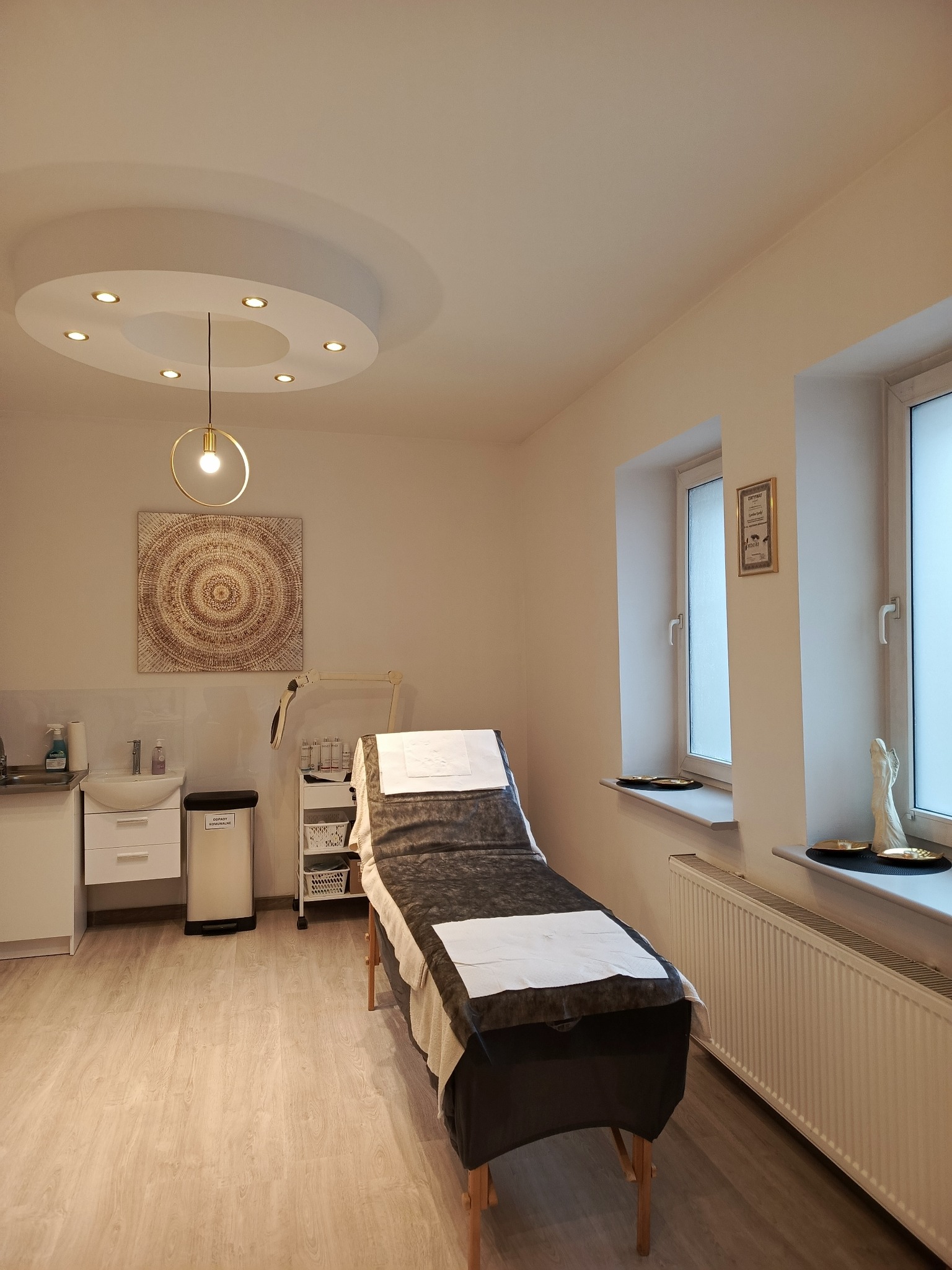 Jasne wnętrze gabinetu z łóżkiem zabiegowym, dekoracyjną lampą i mandalą na ścianie, w tle widoczny umywalnik i okno z dekoracjami.