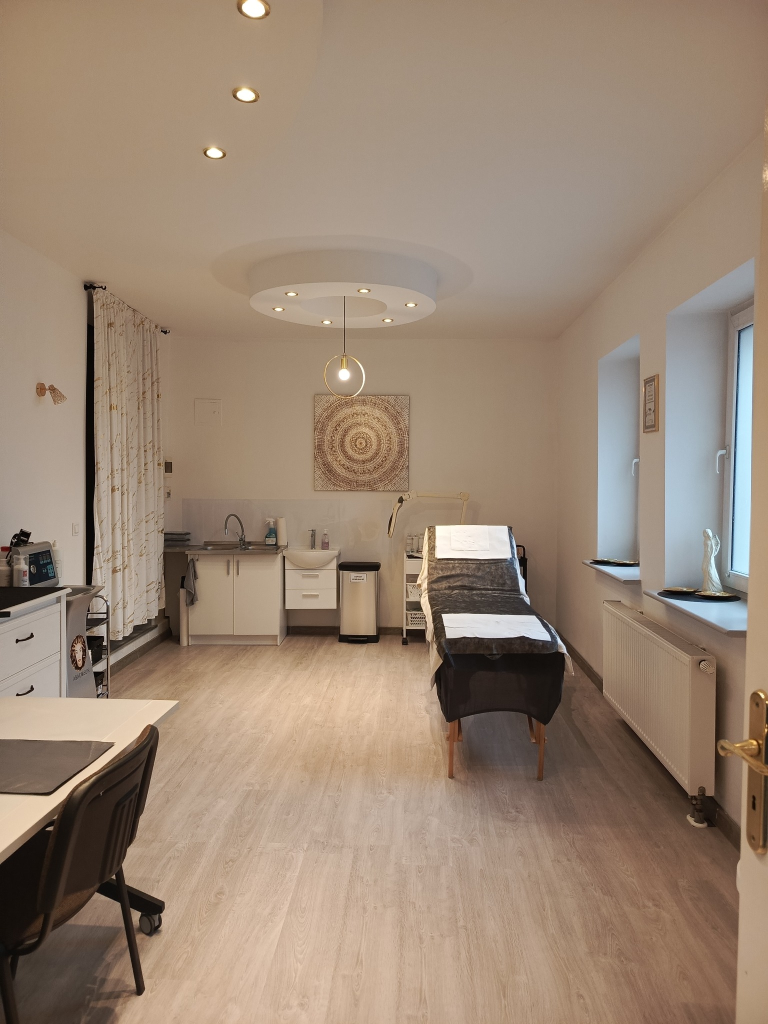 Jasne wnętrze gabinetu kosmetycznego z łóżkiem zabiegowym przykrytym czarną narzutą, umywalką i sprzętem kosmetycznym, oświetlone lampami sufitowymi i naturalnym światłem z okien.