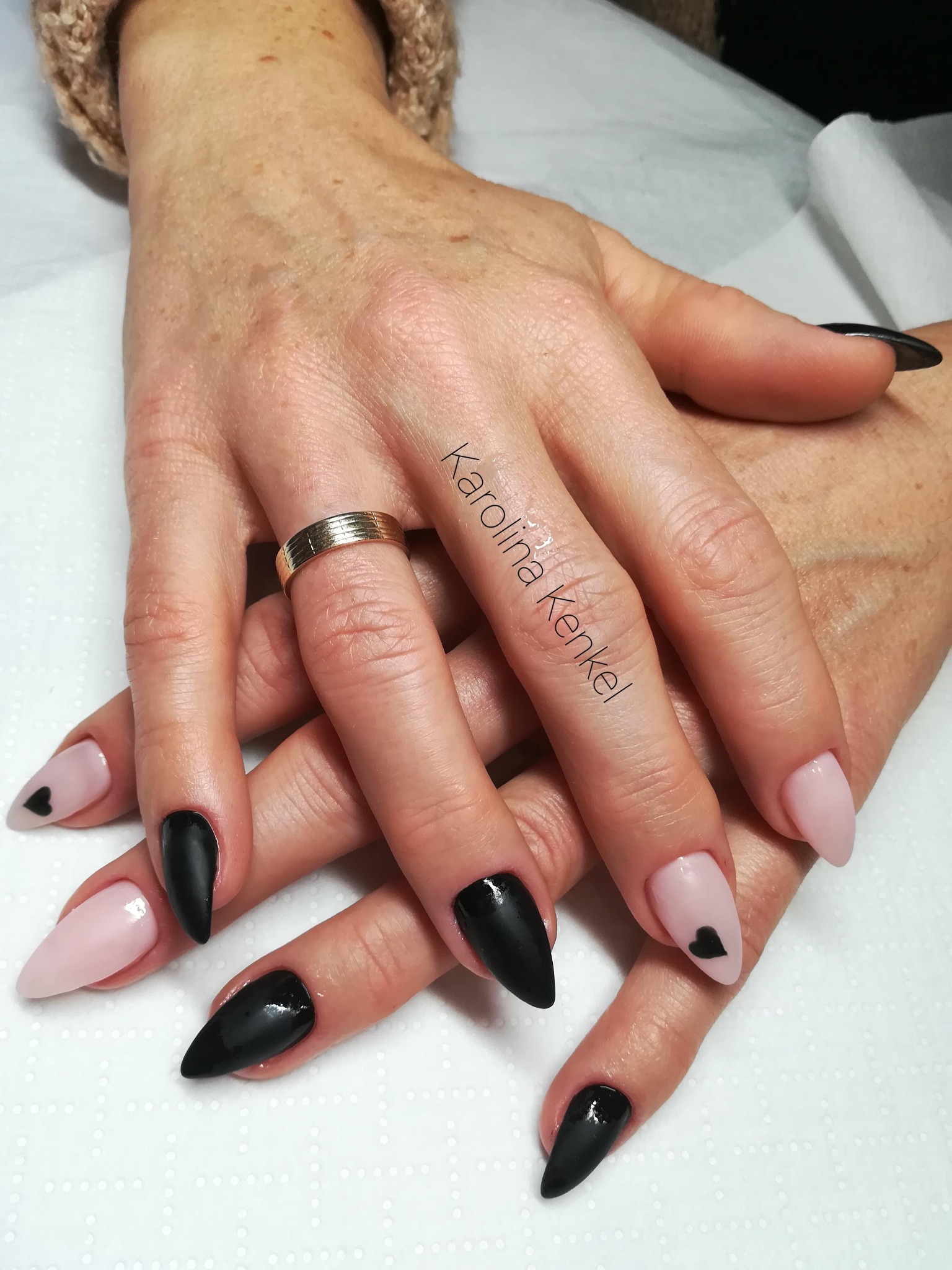 Dłonie kobiety z czarno-różowym manicure hybrydowym w kształcie szpica, z czarnymi serduszkami na niektórych paznokciach i złotym pierścionkiem na palcu.