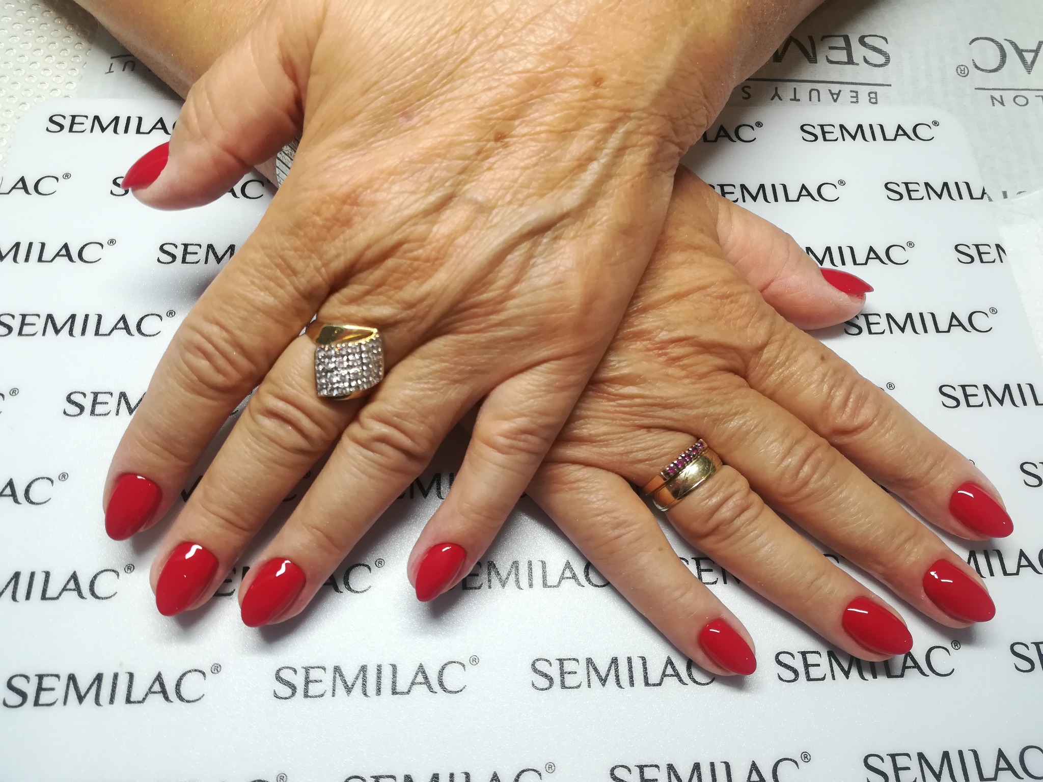 Dłonie kobiety z czerwonymi, błyszczącymi paznokciami na tle podkładki z logo Semilac, widoczne złote pierścionki na palcach.