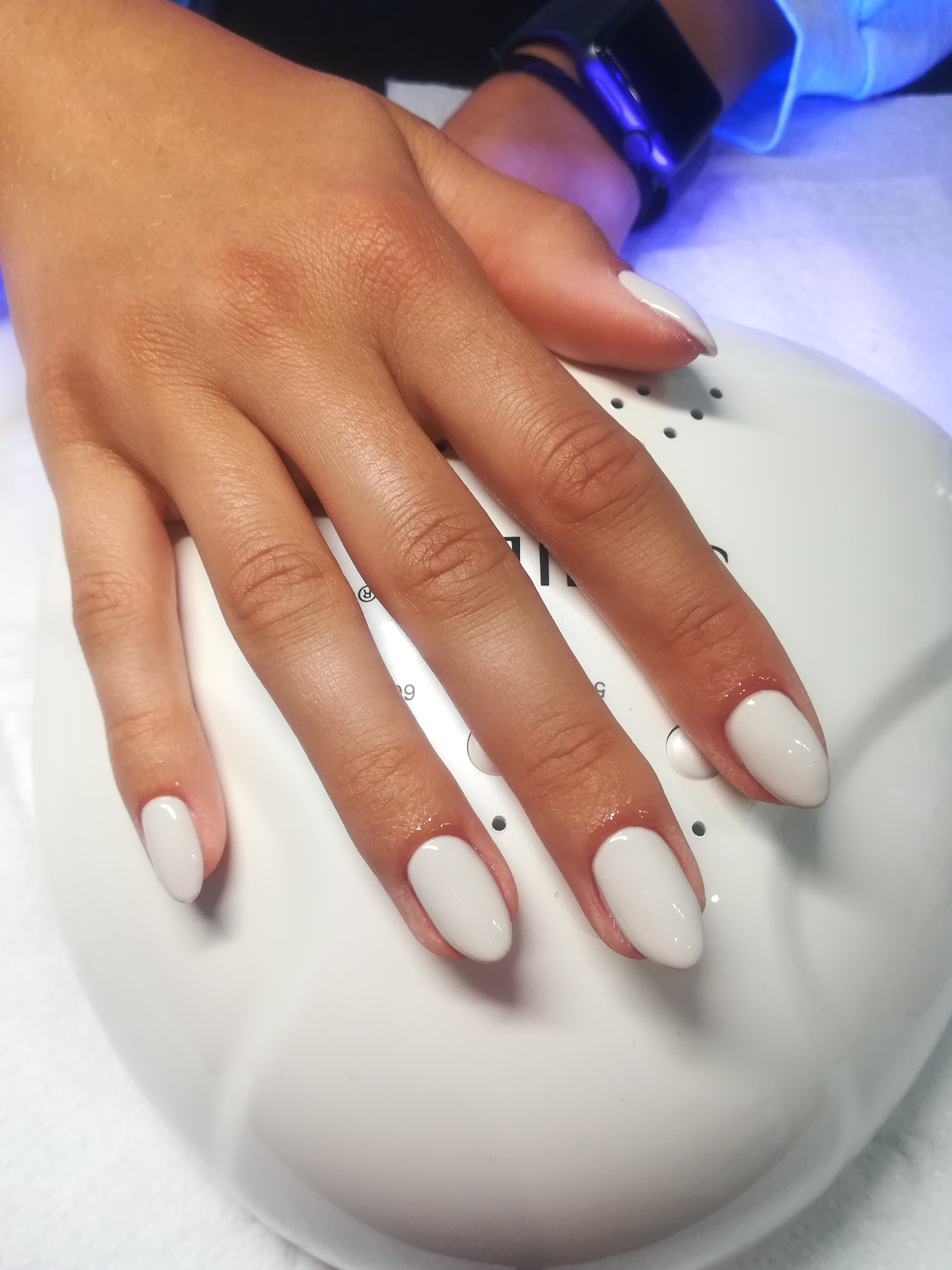 Dłoń z świeżo pomalowanymi, owalnymi paznokciami w odcieniu jasnej szarości, utwardzane w lampie UV, widoczny smartwatch na nadgarstku.