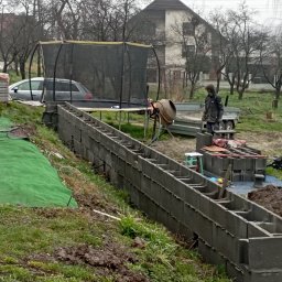 PAWEŁ MACHURA - Budowa muru oporowego z szarych betonowych bloczków na nierównym terenie działki, widoczna betoniarka, taczka, stosy kostki brukowej i trawa.