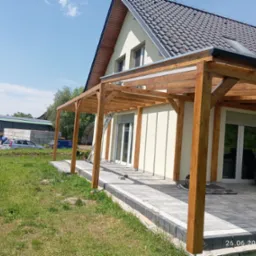 Drewniana pergola z transparentnym zadaszeniem, przylegająca do jasnego budynku z szarą dachówką. Widoczny fragment tarasu wyłożonego kostką brukową i zielony trawnik.