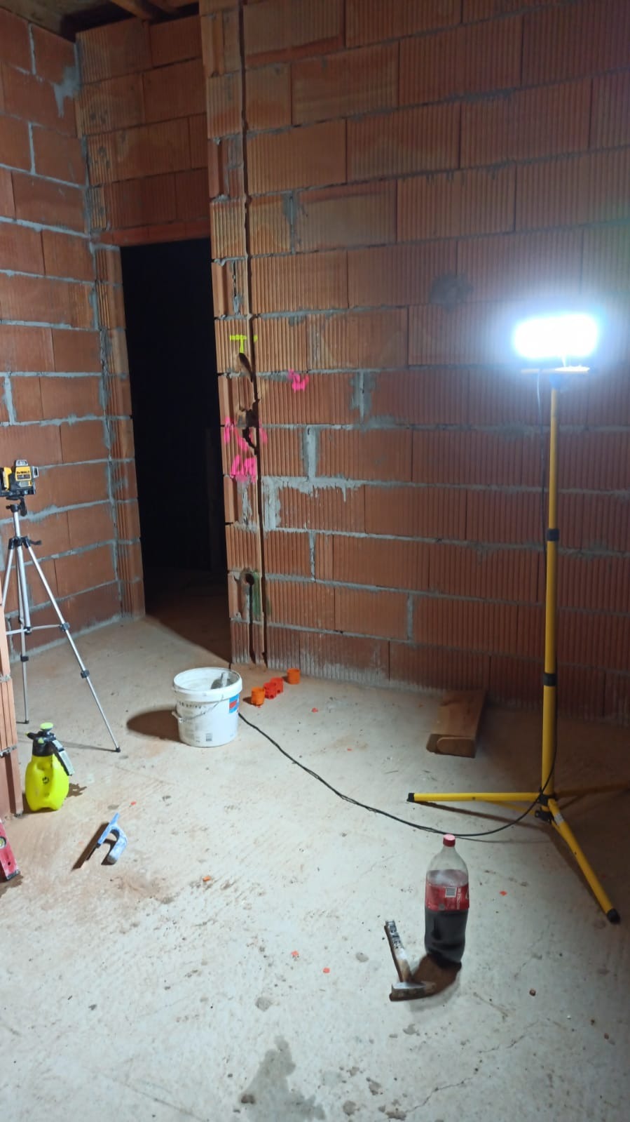 Surowy stan wnętrza z czerwonej cegły dziurawki, podświetlony lampą budowlaną, z widocznym laserowym niwelatorem na statywie, wiadrem z zaprawą, narzędziami i zaznaczeniami na ścianie, ukazujący...