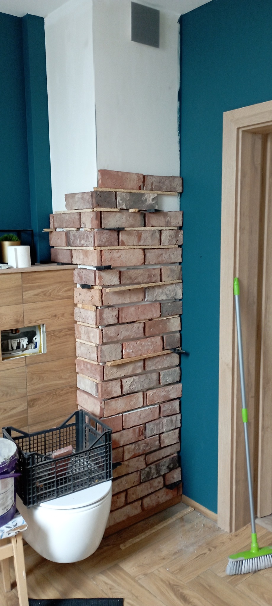 Montaż dekoracyjnej ściany z cegły klinkierowej w łazience, widoczne drewniane listwy dystansowe, ściana pomalowana na turkusowo, w tle toaleta i podłoga z paneli.