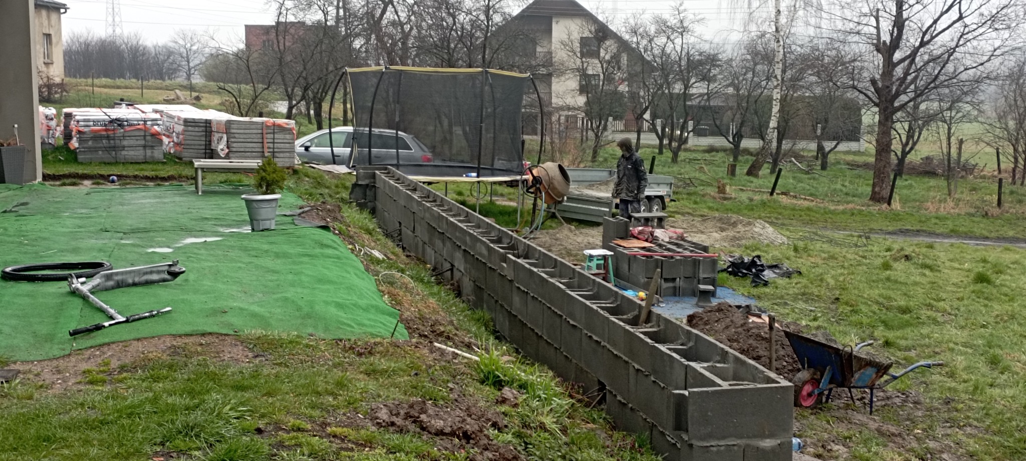 Budowa muru oporowego z szarych betonowych bloczków na nierównym terenie działki, widoczna betoniarka, taczka, stosy kostki brukowej i trawa.
