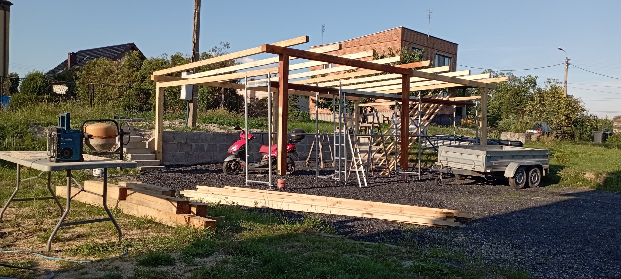 Konstrukcja drewnianej wiaty w trakcie budowy, widoczne rusztowania, przyczepka samochodowa, skuter oraz betoniarka z agregatem prądotwórczym.