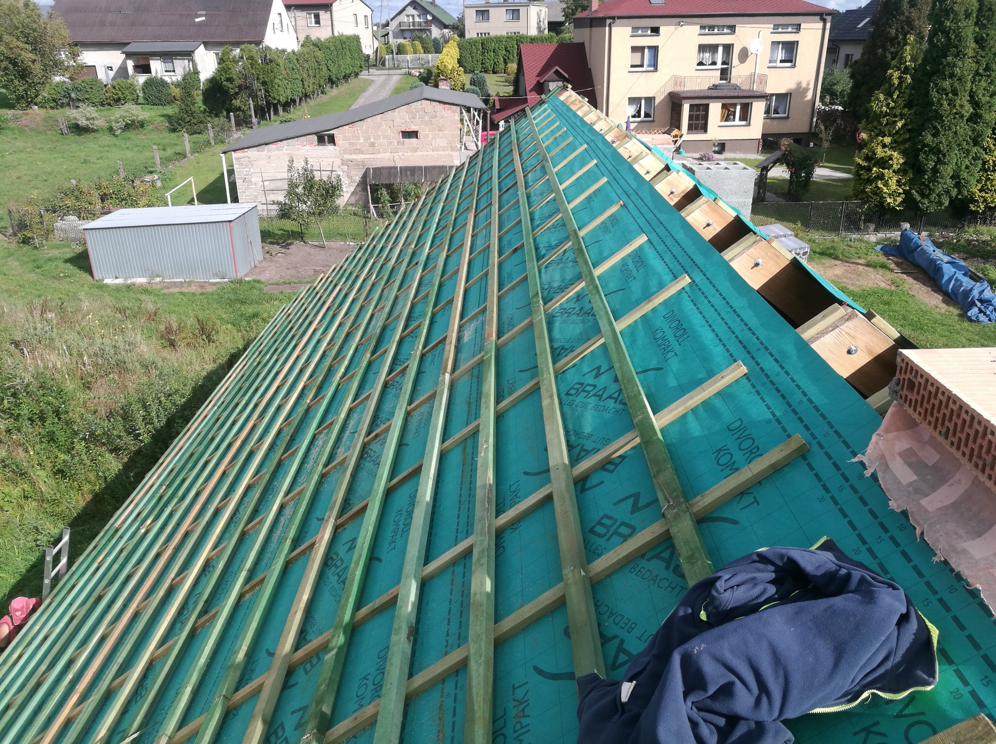 Konstrukcja dachu z drewnianych łat pokrytych zieloną membraną hydroizolacyjną Braas Divoroll Kompakt, widok z góry na tle zabudowań mieszkalnych i zieleni.