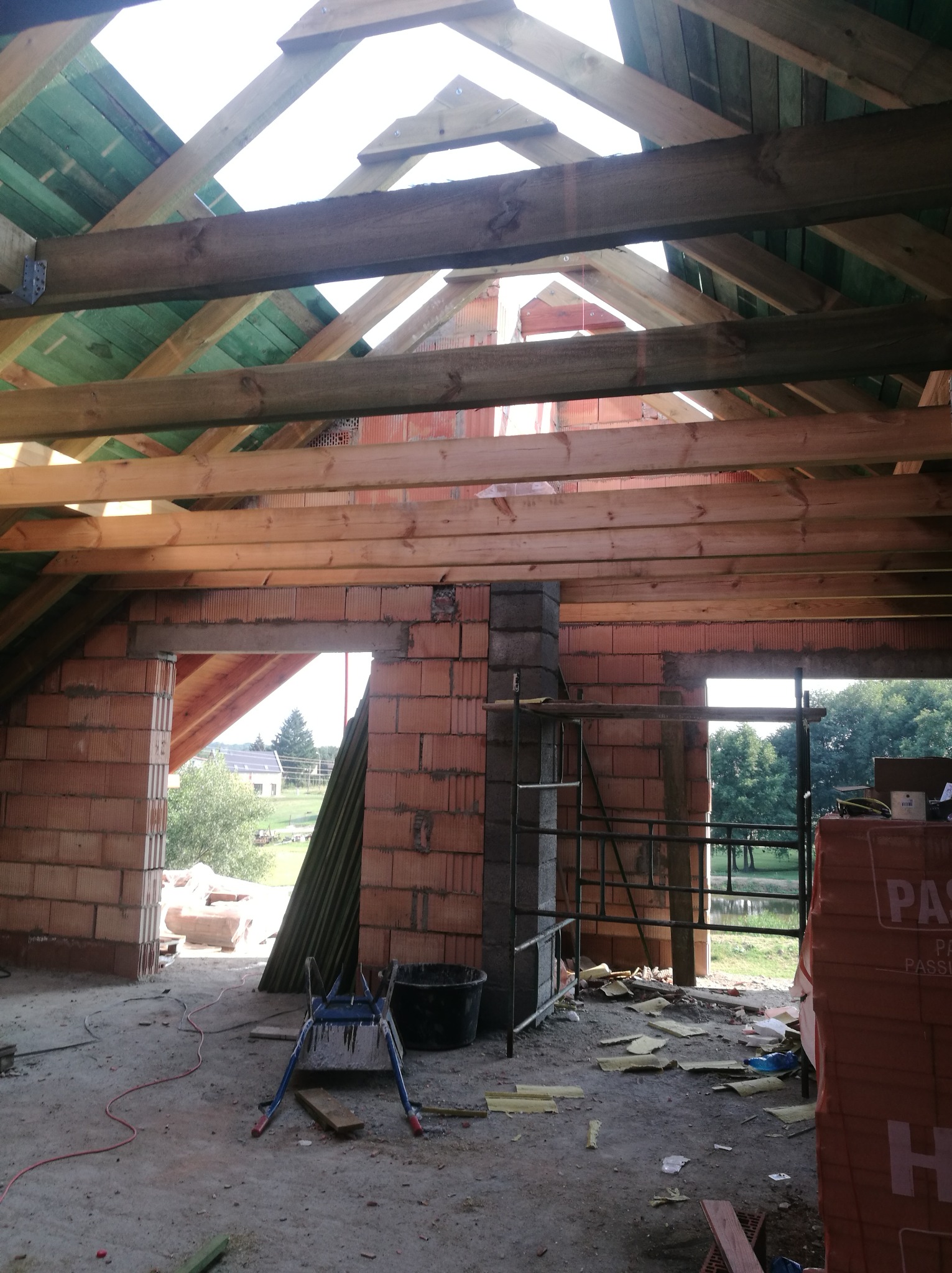 Wnętrze w trakcie budowy: surowa konstrukcja dachu z drewnianych belek i zielonej membrany, ściany z czerwonej cegły dziurawki, widoczne otwory okienne i drzwiowe, na podłodze resztki materiałów...