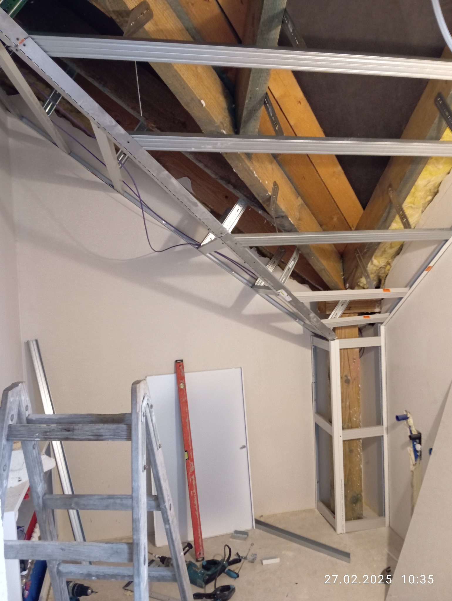 Stelaż sufitowy z profili metalowych w trakcie montażu, widoczne drewniane belki stropowe, drabina malarska i narzędzia na podłodze.