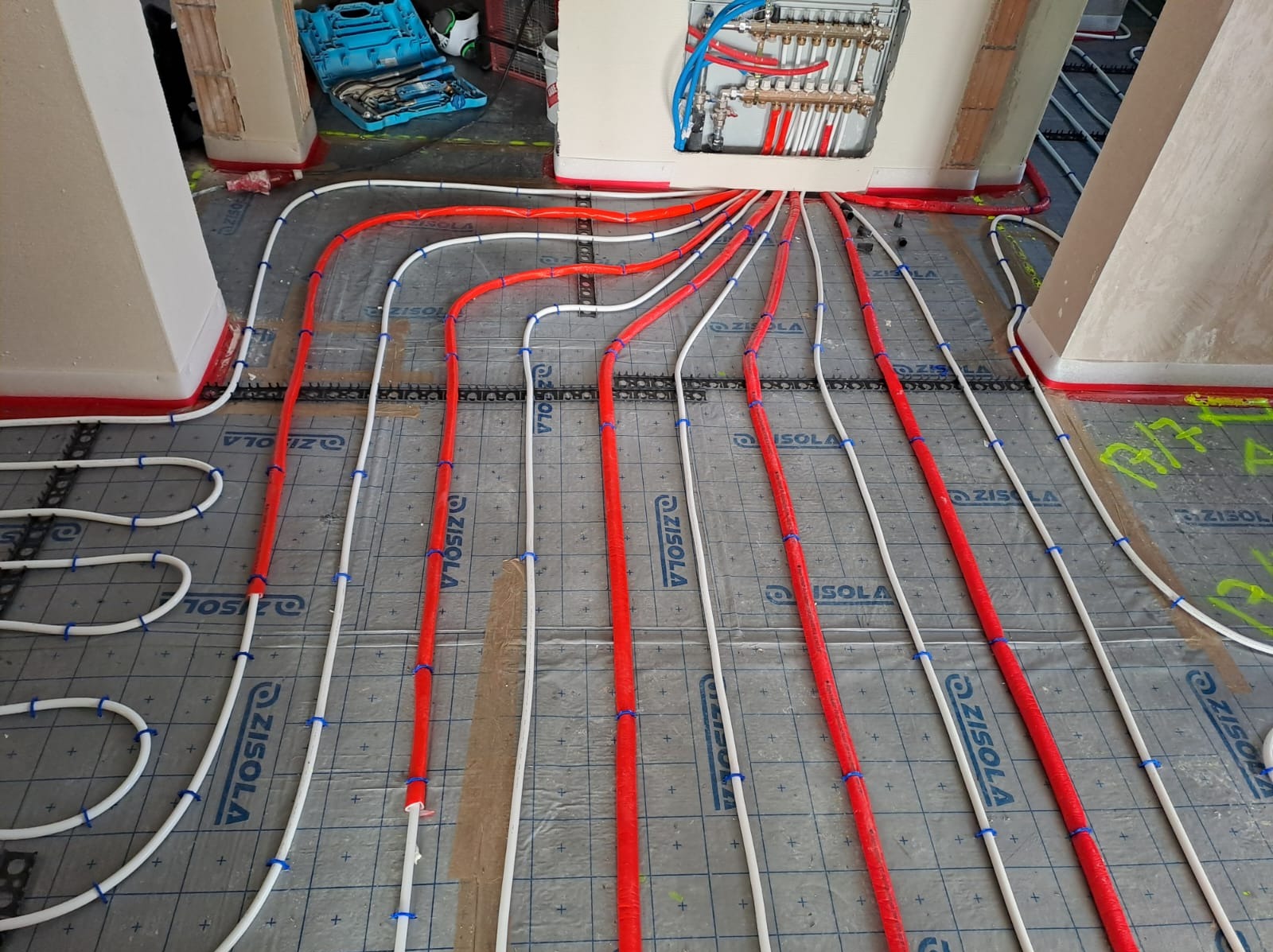 Montaż ogrzewania podłogowego: czerwone i białe rury układane na folii izolacyjnej Zisola, przymocowane niebieskimi uchwytami, z widocznym rozdzielaczem w ścianie i skrzynką z narzędziami w tle.