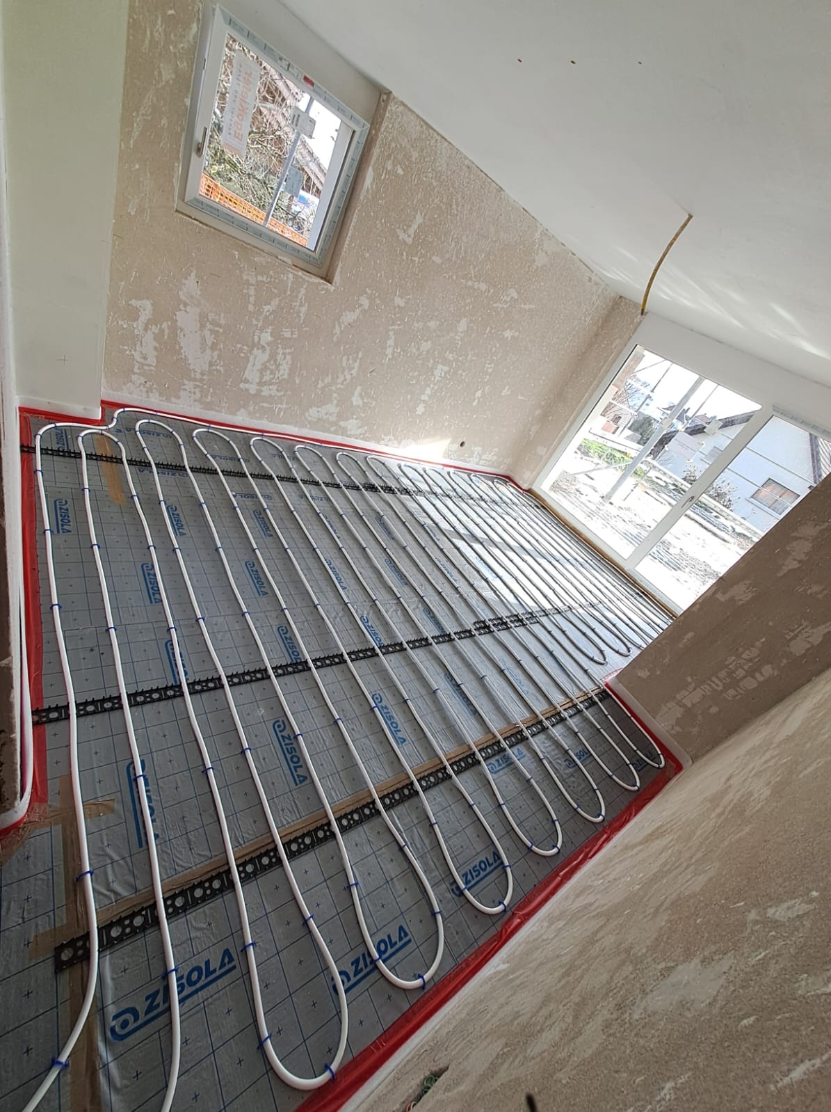 Montaż ogrzewania podłogowego w pomieszczeniu z przygotowanymi ścianami, widoczne rury grzewcze ułożone na izolacji z folią aluminiową i uchwytami mocującymi, okno w tle.