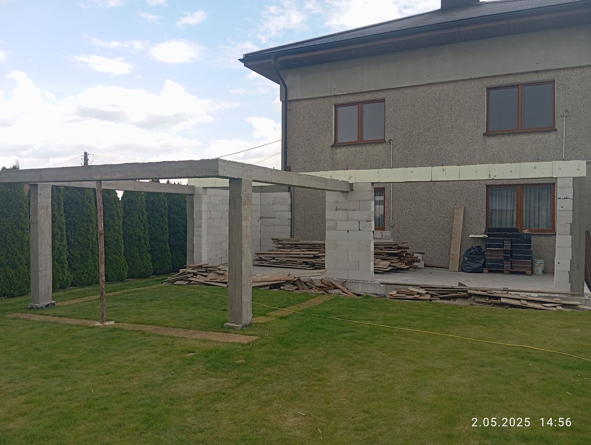 Budowa tarasu przy domu: betonowe słupy i ściany z bloczków, przygotowane deski, zielony trawnik. Widok na elewację budynku z oknami. Etap prac w toku.